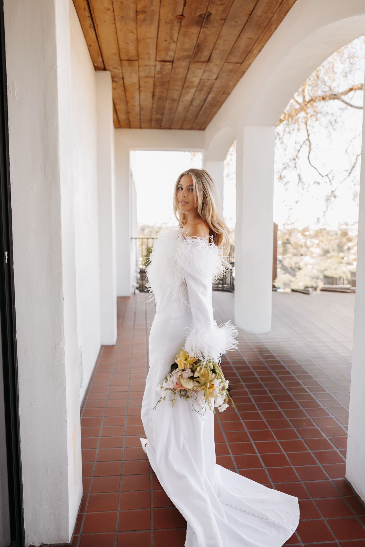 A person in a flowing white wedding dress walks through a Spanish colonial style covered walkway with terracotta tiles.