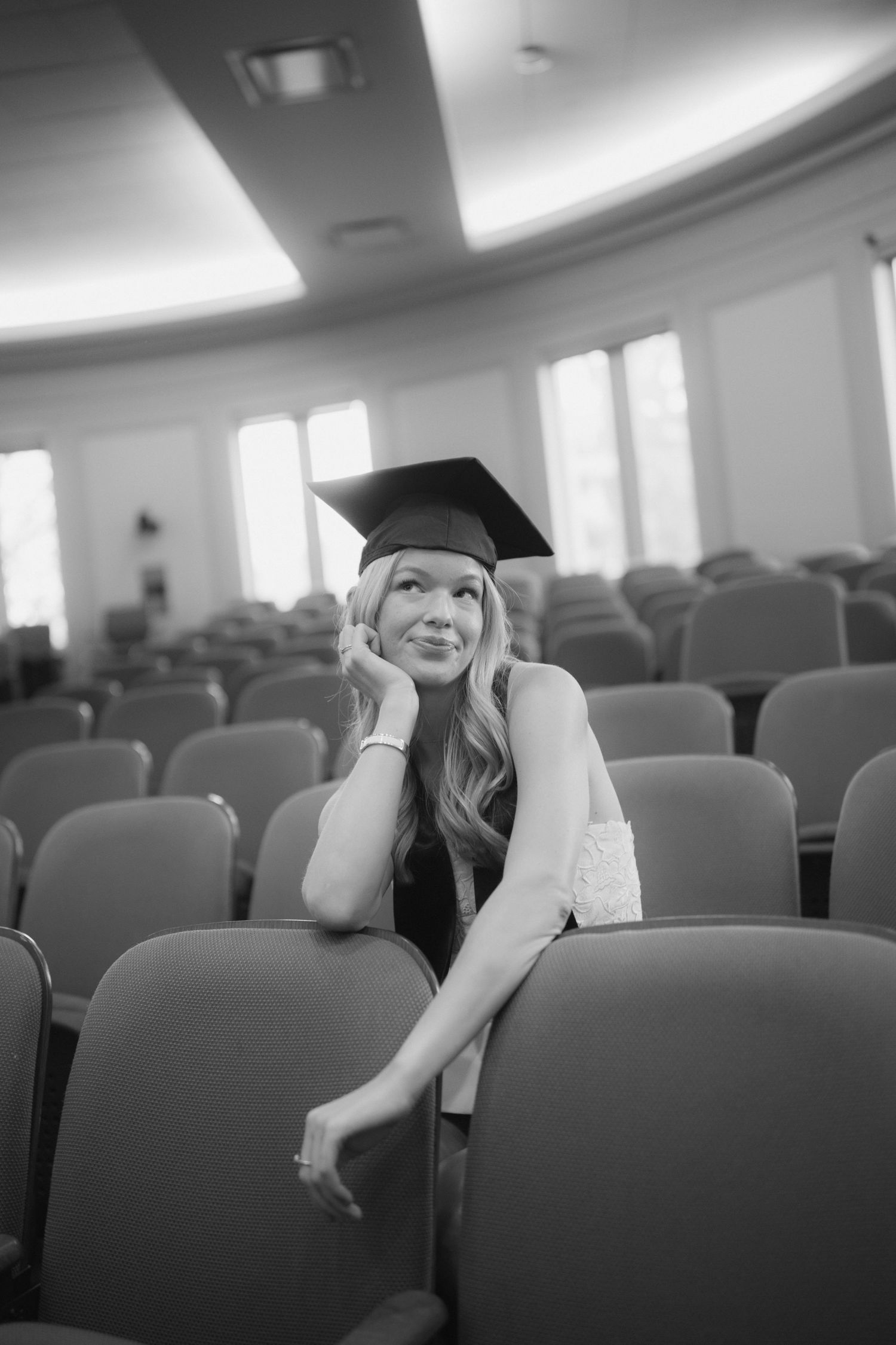 A graduate in cap and gown sits thoughtfully in an empty auditorium with rows of chairs.