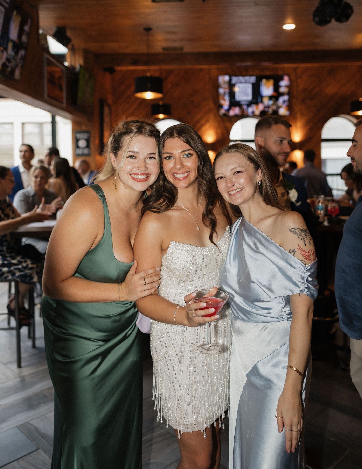 A group of friends in formal attire share joyful moments at what appears to be a wedding reception or celebration.