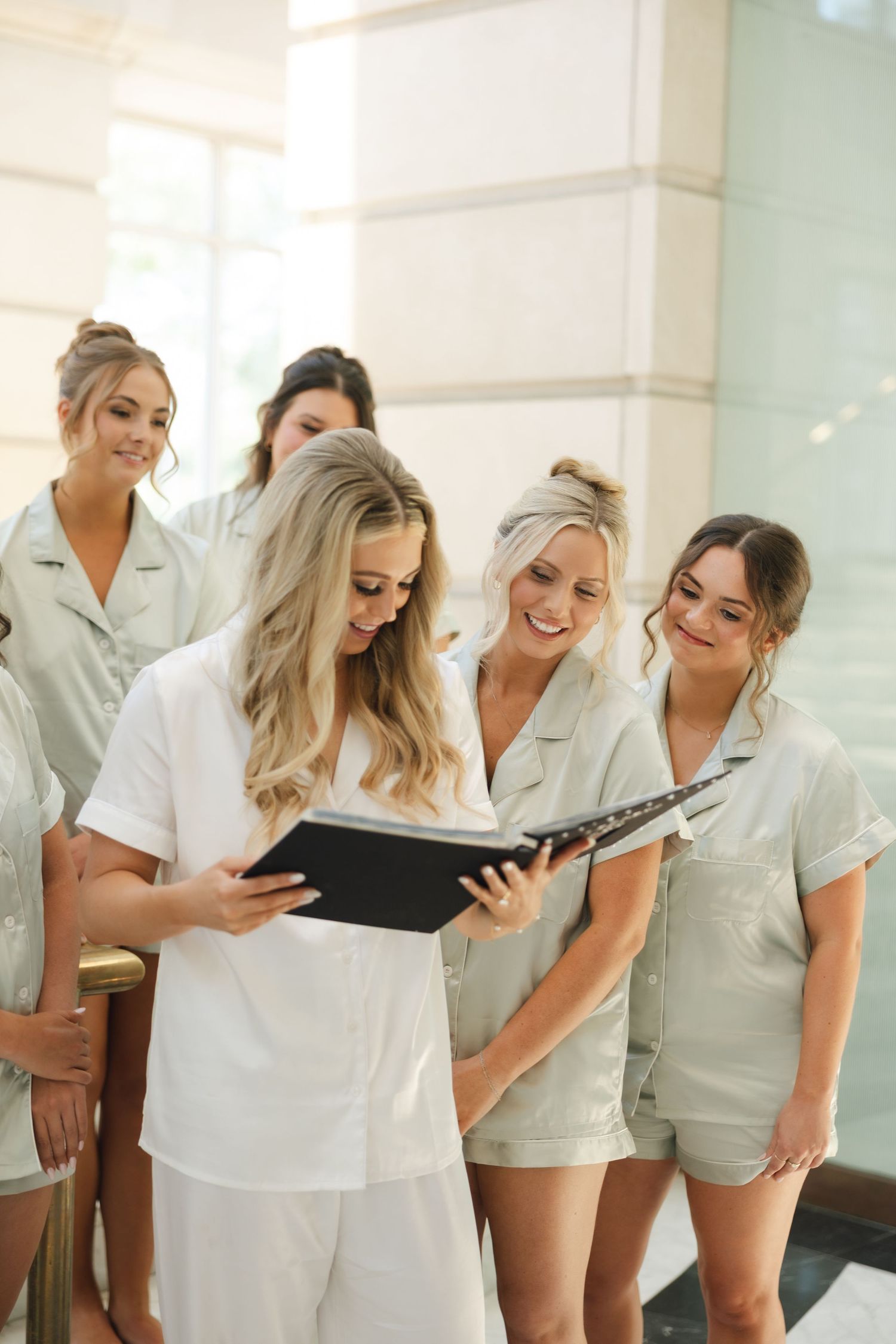 A group of bridesmaids in white robes laugh together while looking at something during wedding preparations.