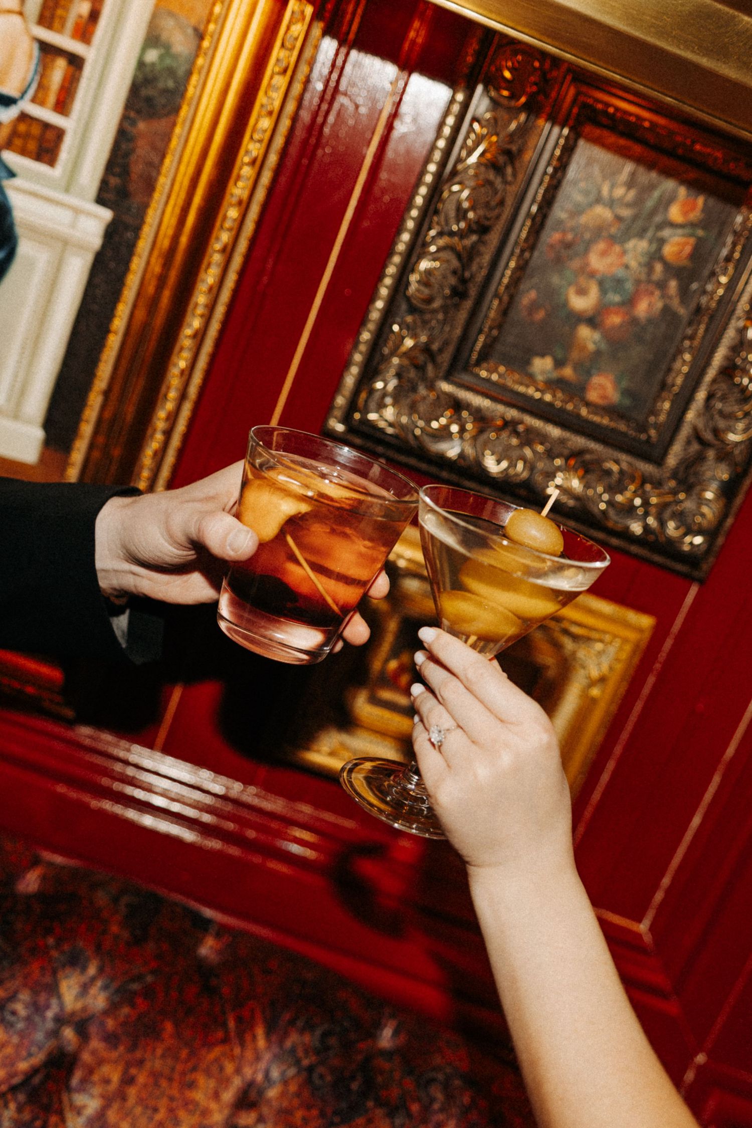 Two hands clink cocktail glasses together in a richly decorated room with red walls and ornate gold frames.