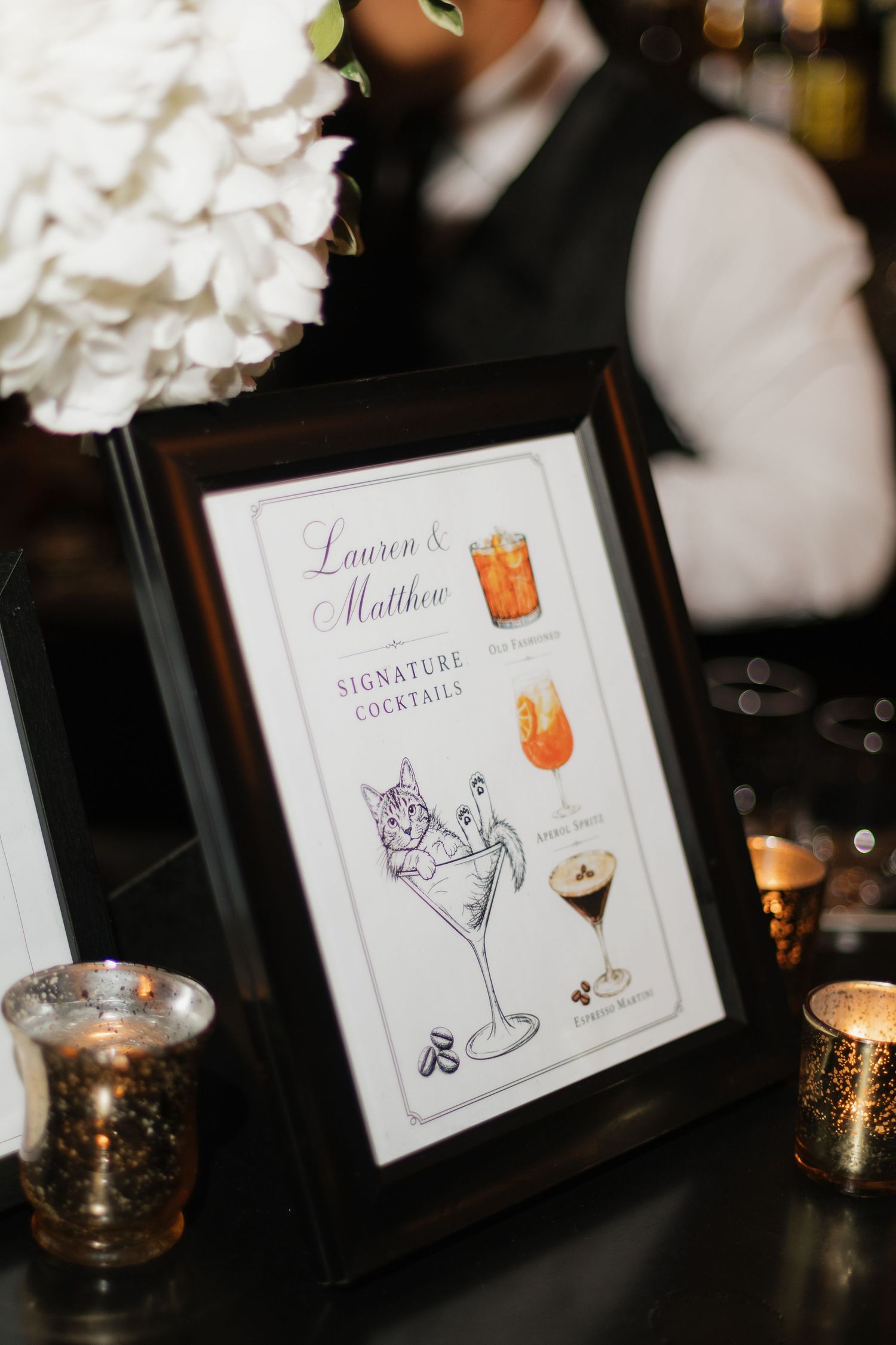 Framed signature cocktails menu for Lauren and Matthew displayed at a wedding bar with candles and white floral decor.