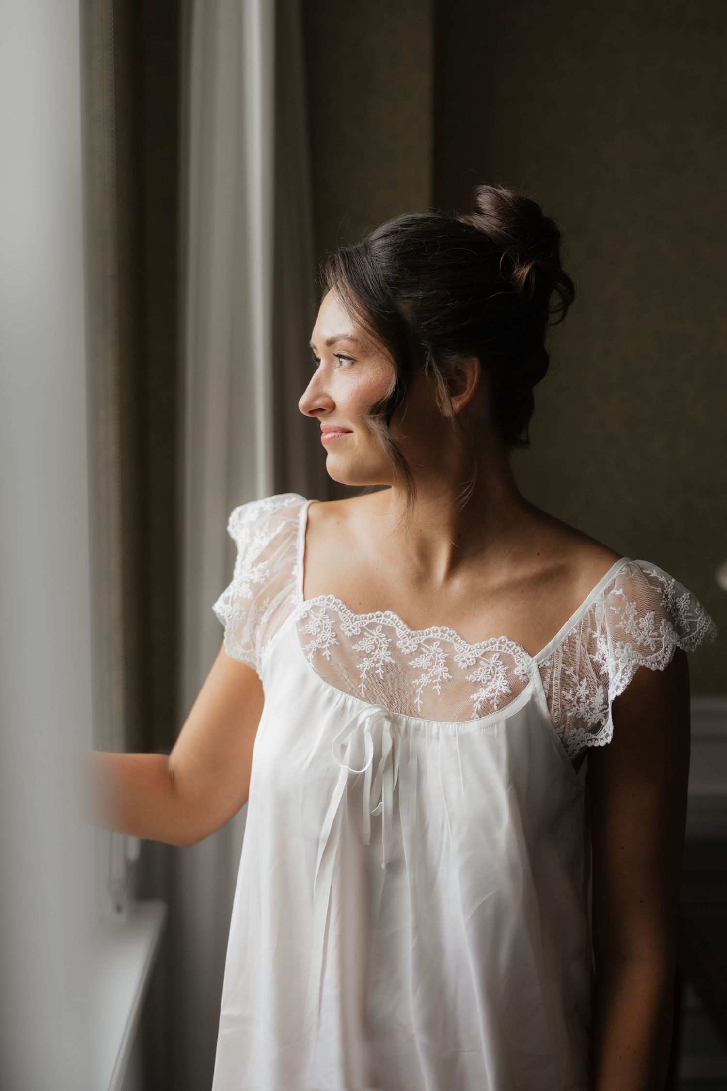 Bride in white lace nightgown gazing out window on wedding morning.