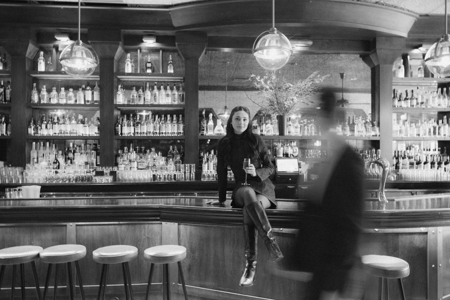 A panoramic black and white view of an elegant bar interior with bottles and glassware displayed on illuminated shelves.