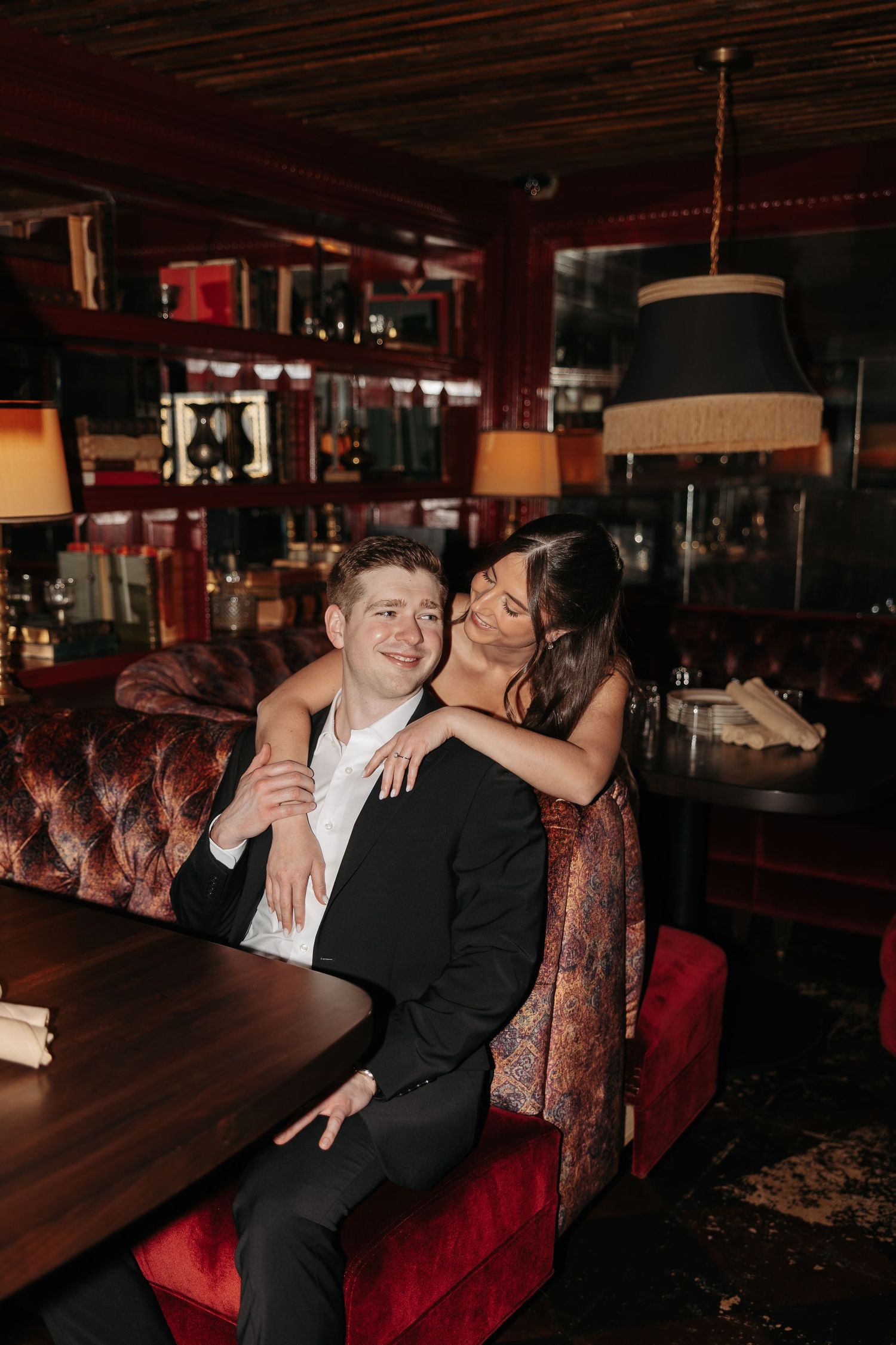 Couple sharing intimate moments on red velvet booth seating in dimly lit restaurant or lounge setting.