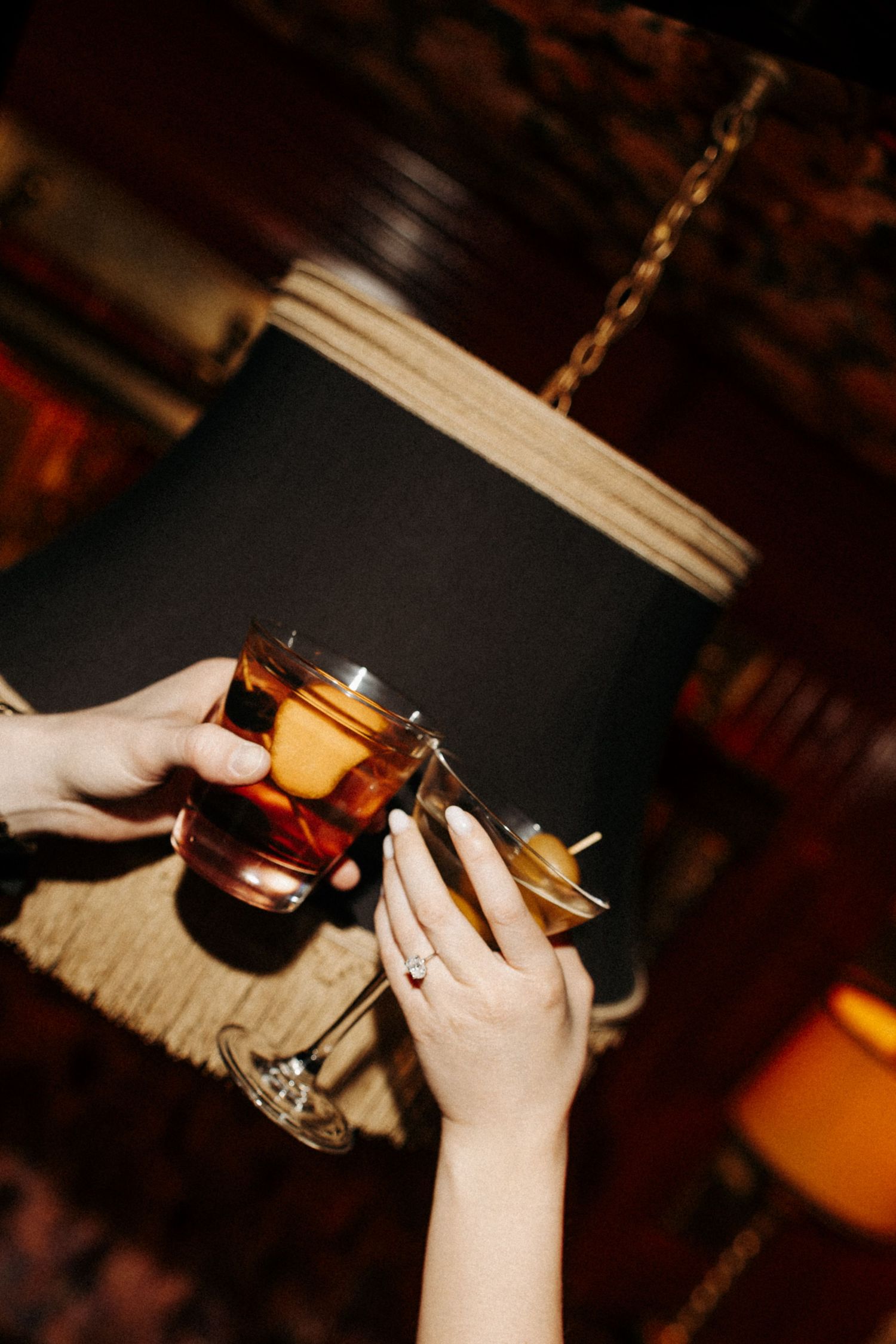 Two hands holding cocktail glasses toasting in a dimly lit bar with warm ambient lighting.