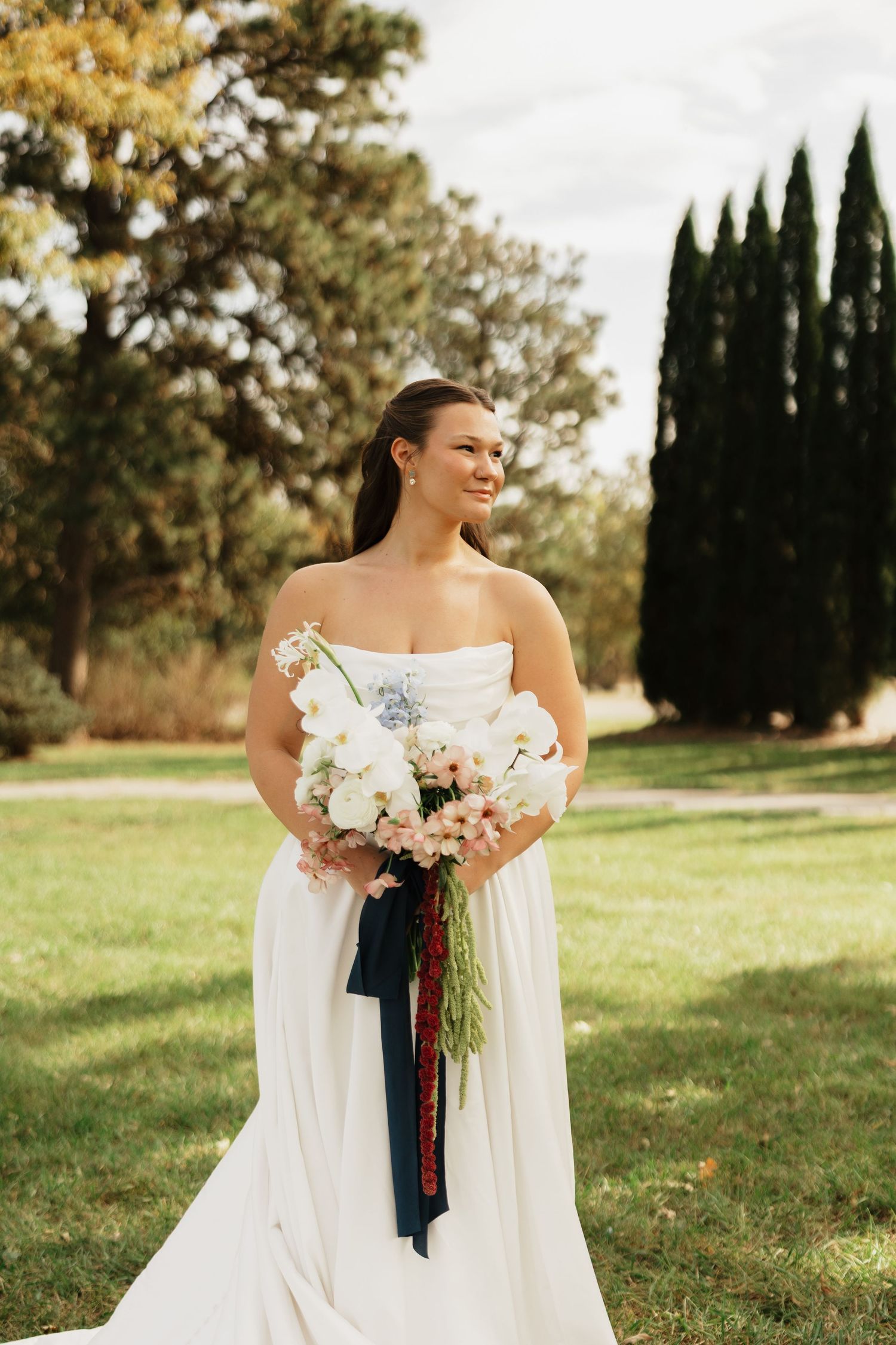 Bride in a strapless white gown holds a pink and white bouquet with navy and red ribbons on a sunny lawn near tall evergreen trees.