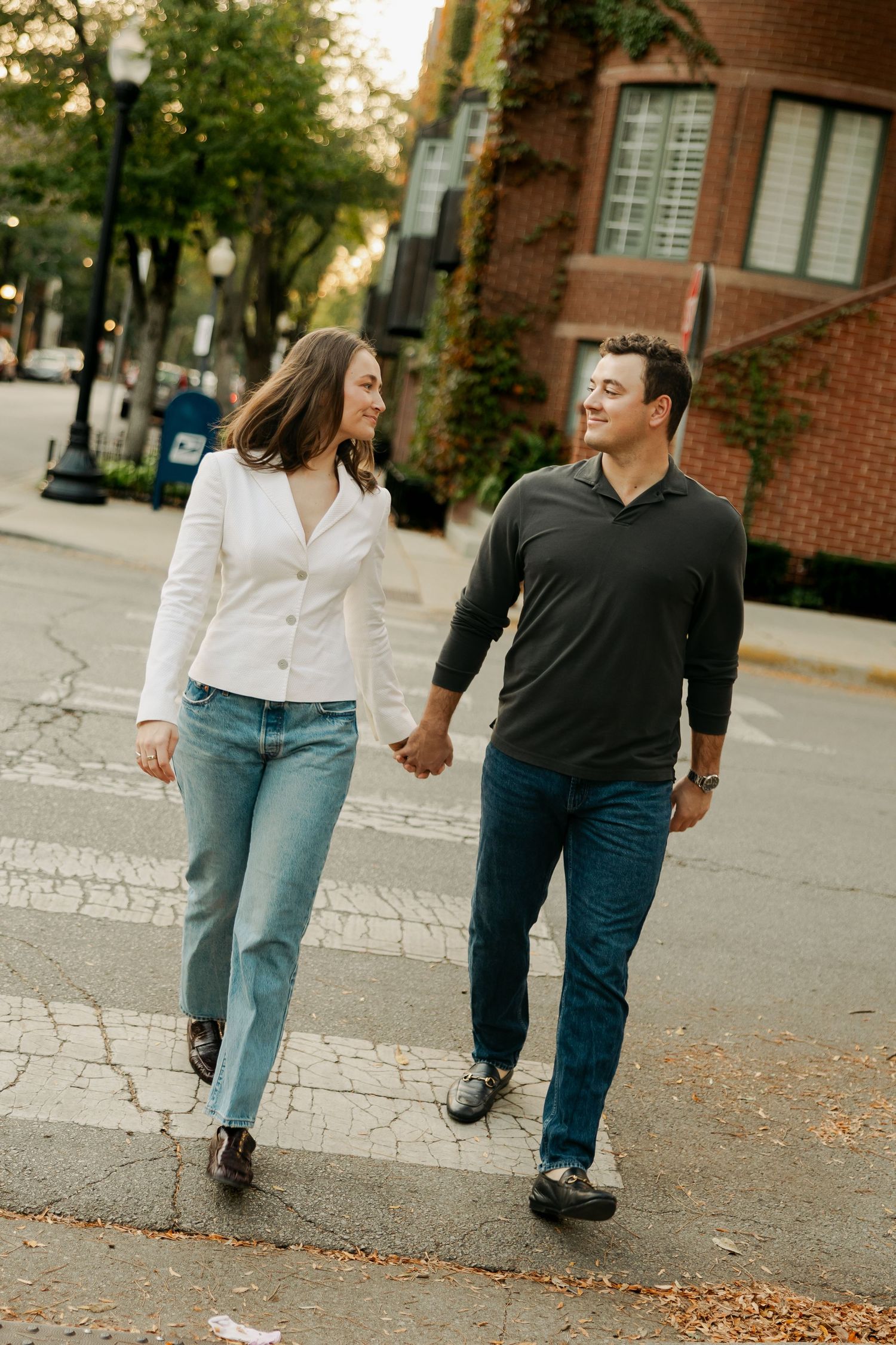 A smiling couple holds hands while walking across a tree-lined city crosswalk, past brick buildings covered in ivy.