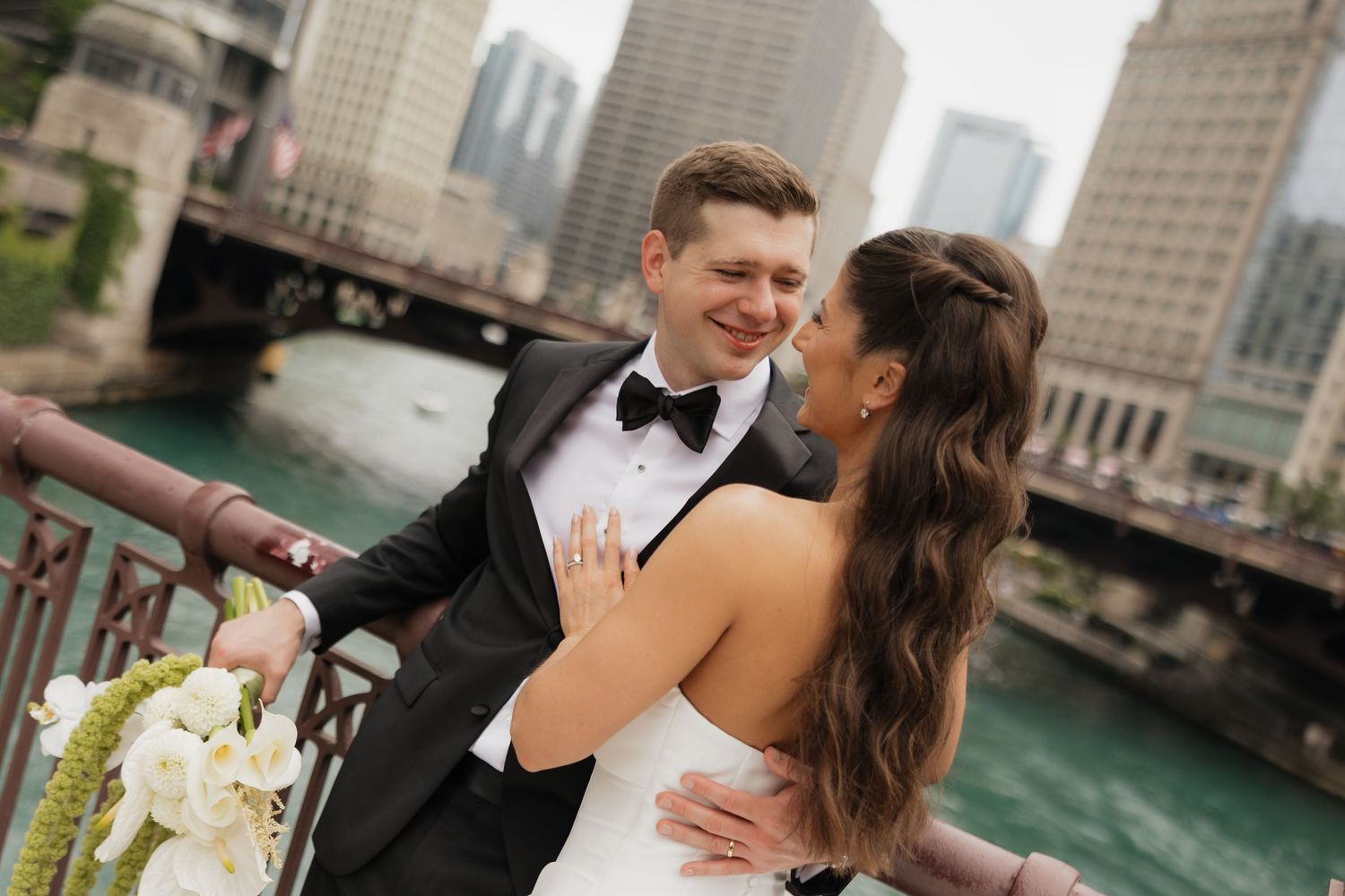 Smiling groom in a black tuxedo and bow tie embraces his bride in a white gown on a riverside bridge with city skyscrapers behind them.