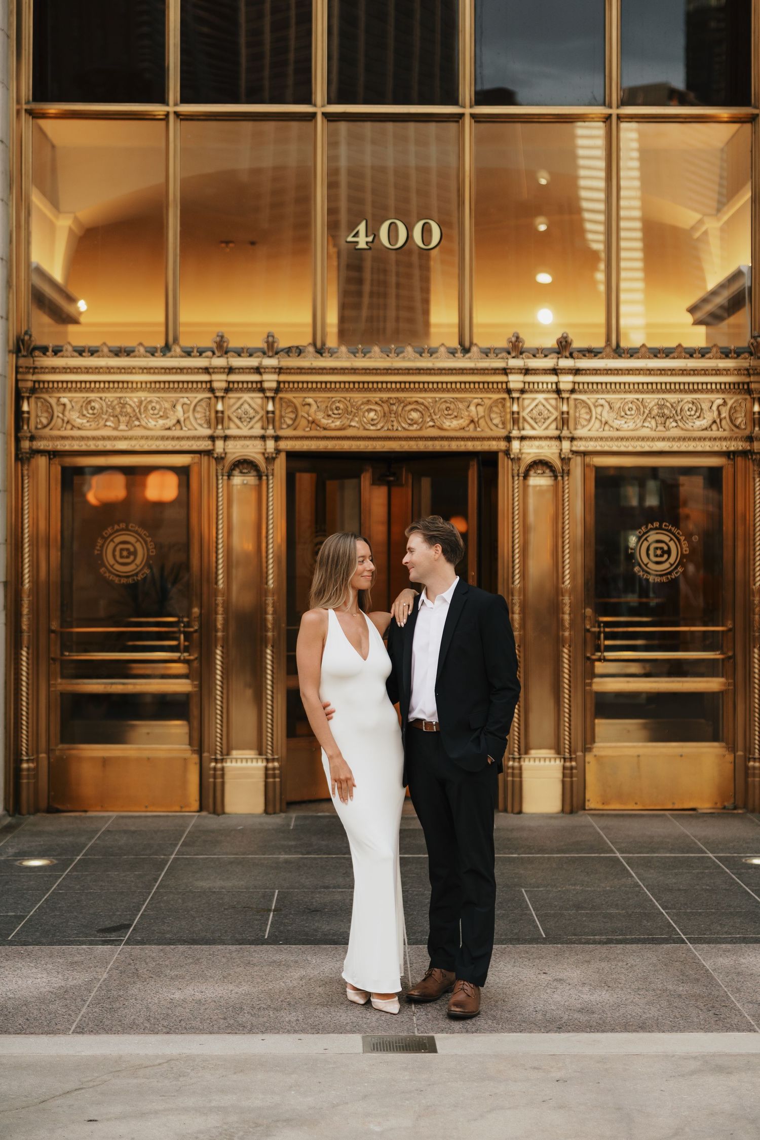 Couple in a white slip dress and black suit embrace and smile in front of ornate golden doors marked 400 for an engagement photo.