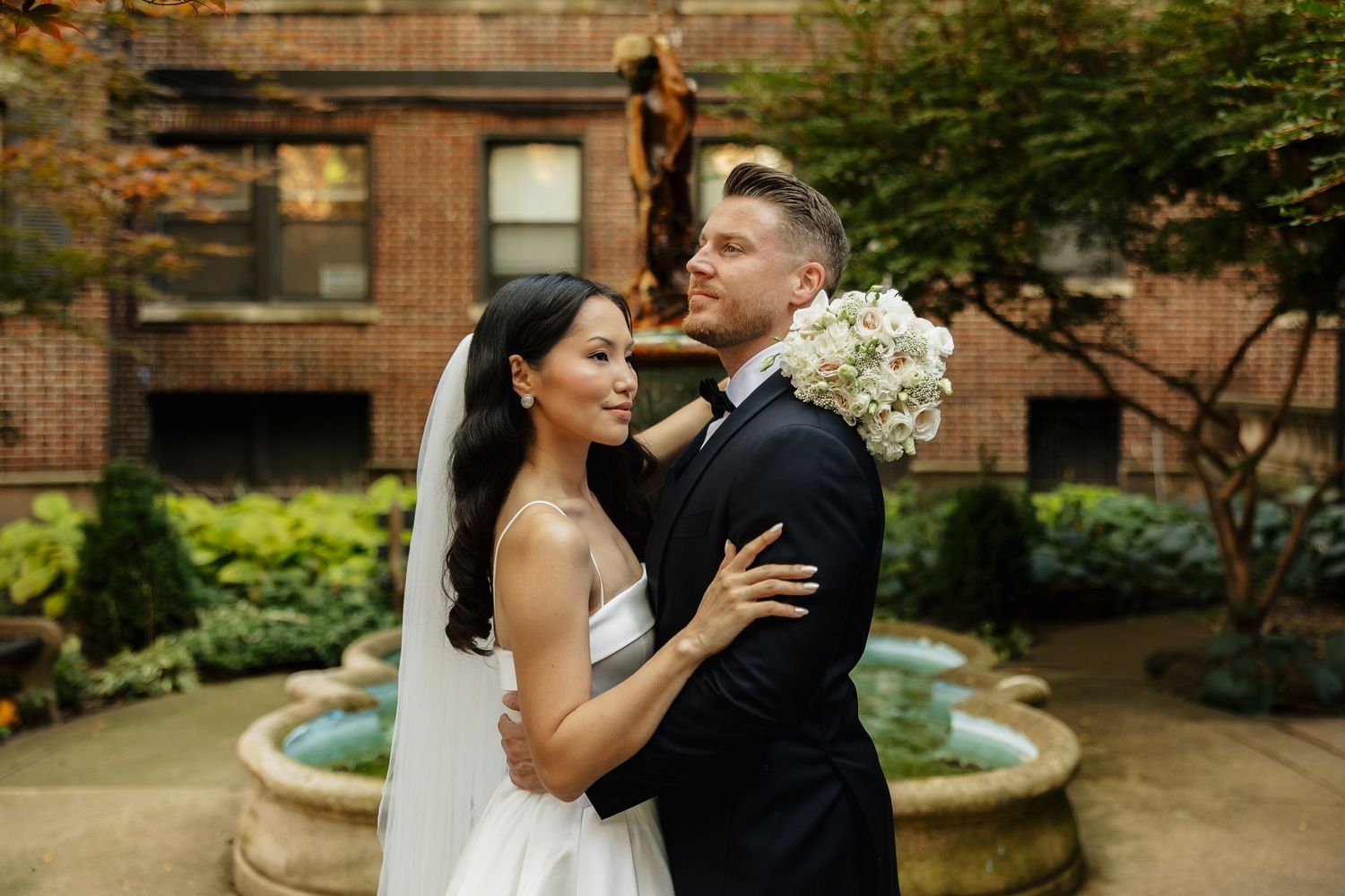 Bride in a white strapless gown and groom in a dark suit embrace in a courtyard garden with a stone fountain and brick building.