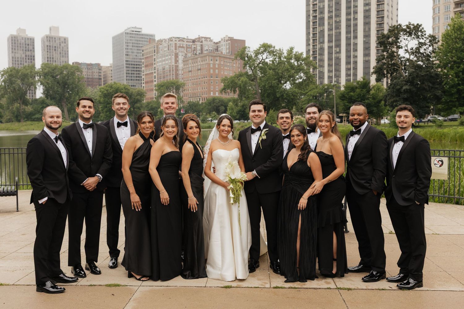 Wedding party in black attire poses with bride and groom outdoors near a city skyline and lush greenery.