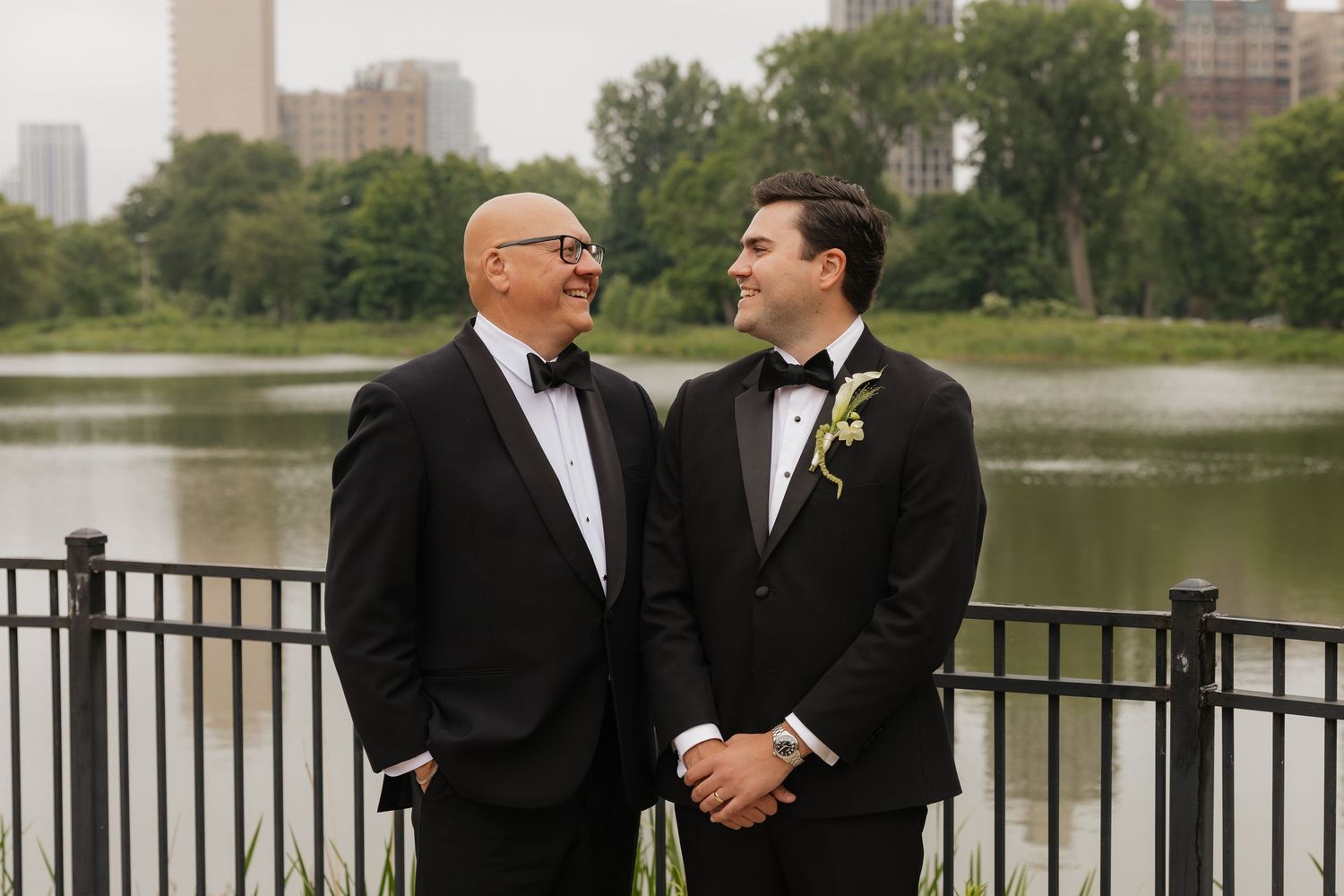 Two grooms in black tuxedos share a tender moment by a lakeside railing with a city skyline backdrop.