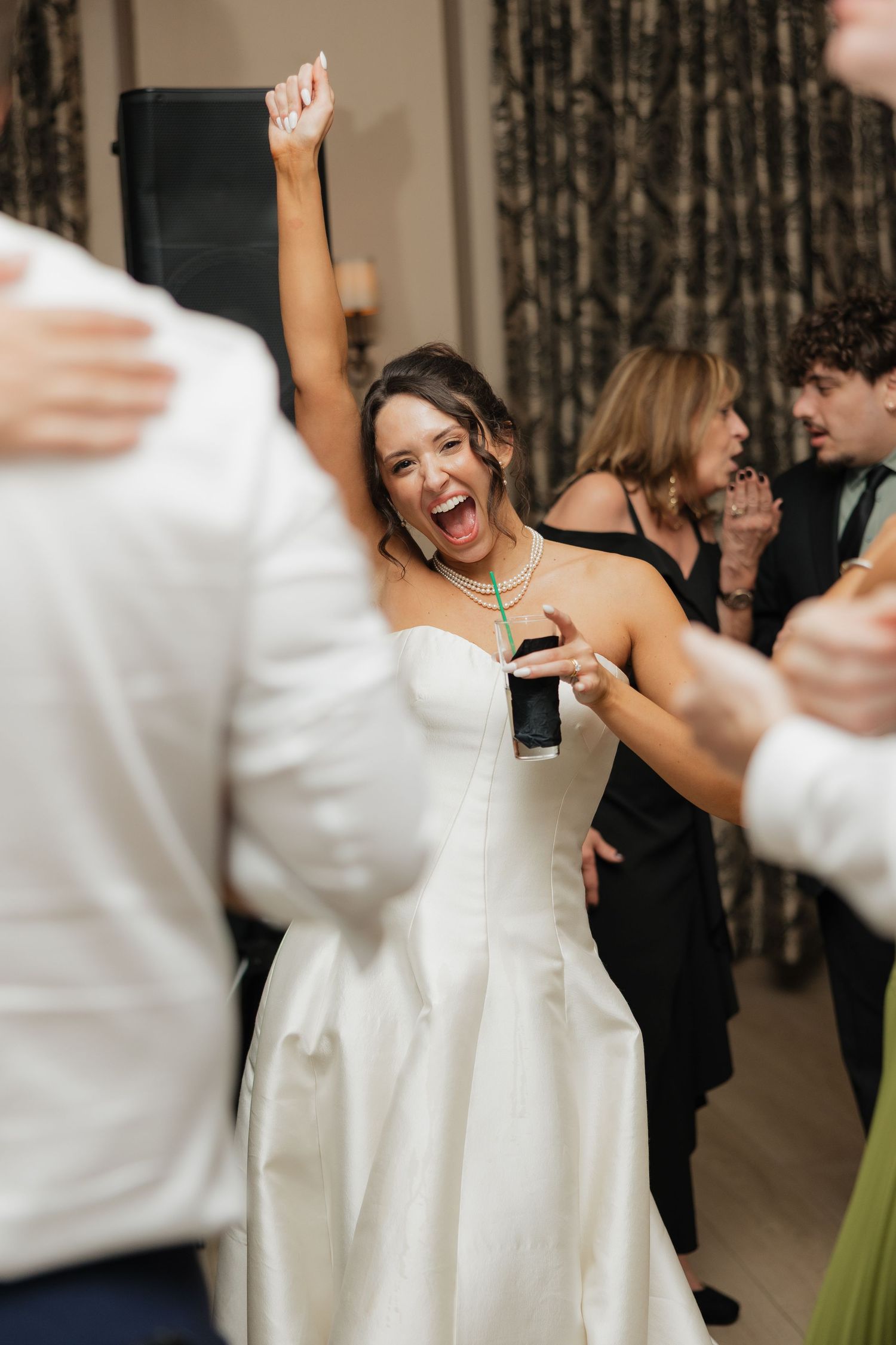 Joyful bride in white strapless gown dancing with raised fist at wedding reception surrounded by guests.