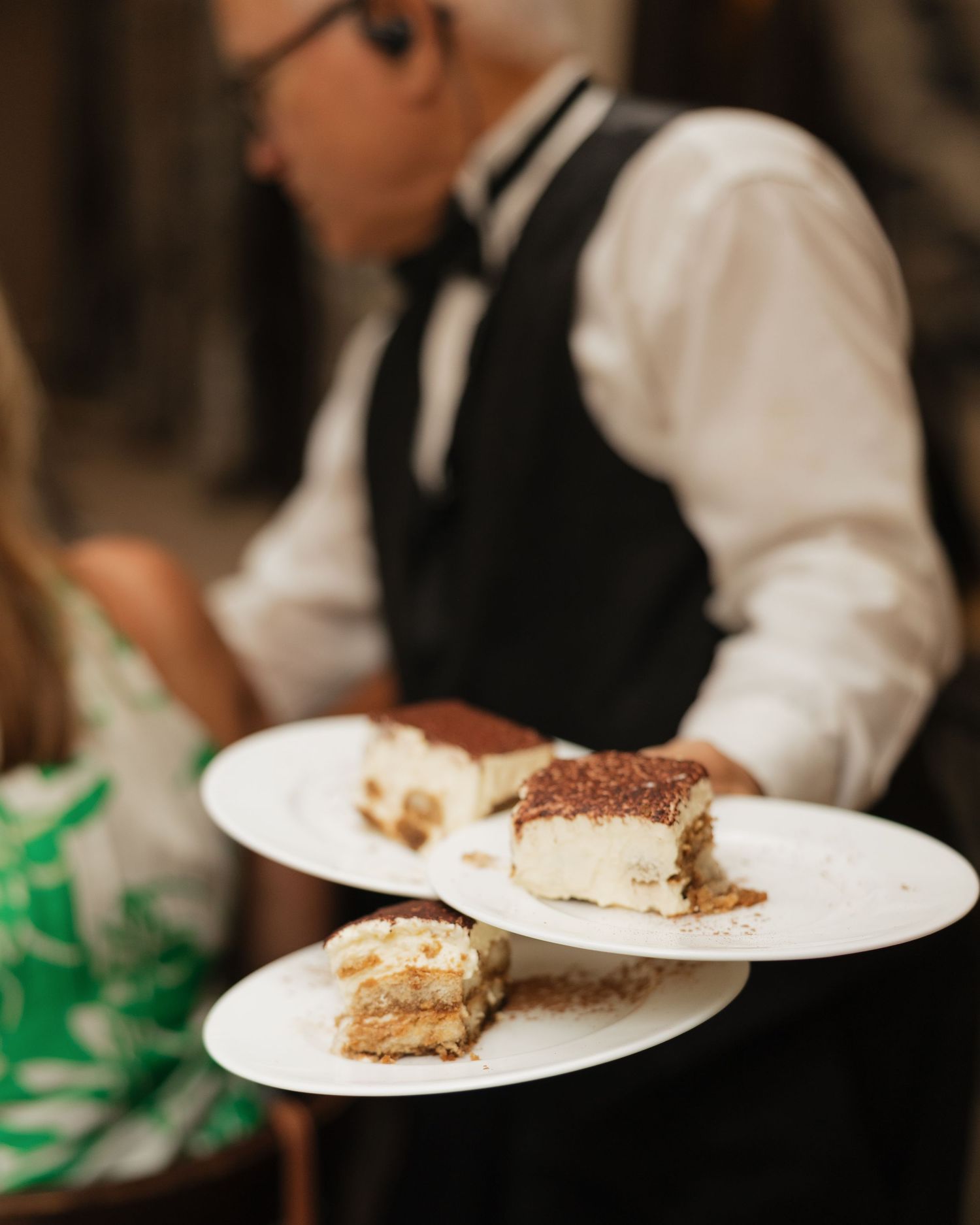 Waiter in vest serving plates of tiramisu dessert at an elegant catered event.