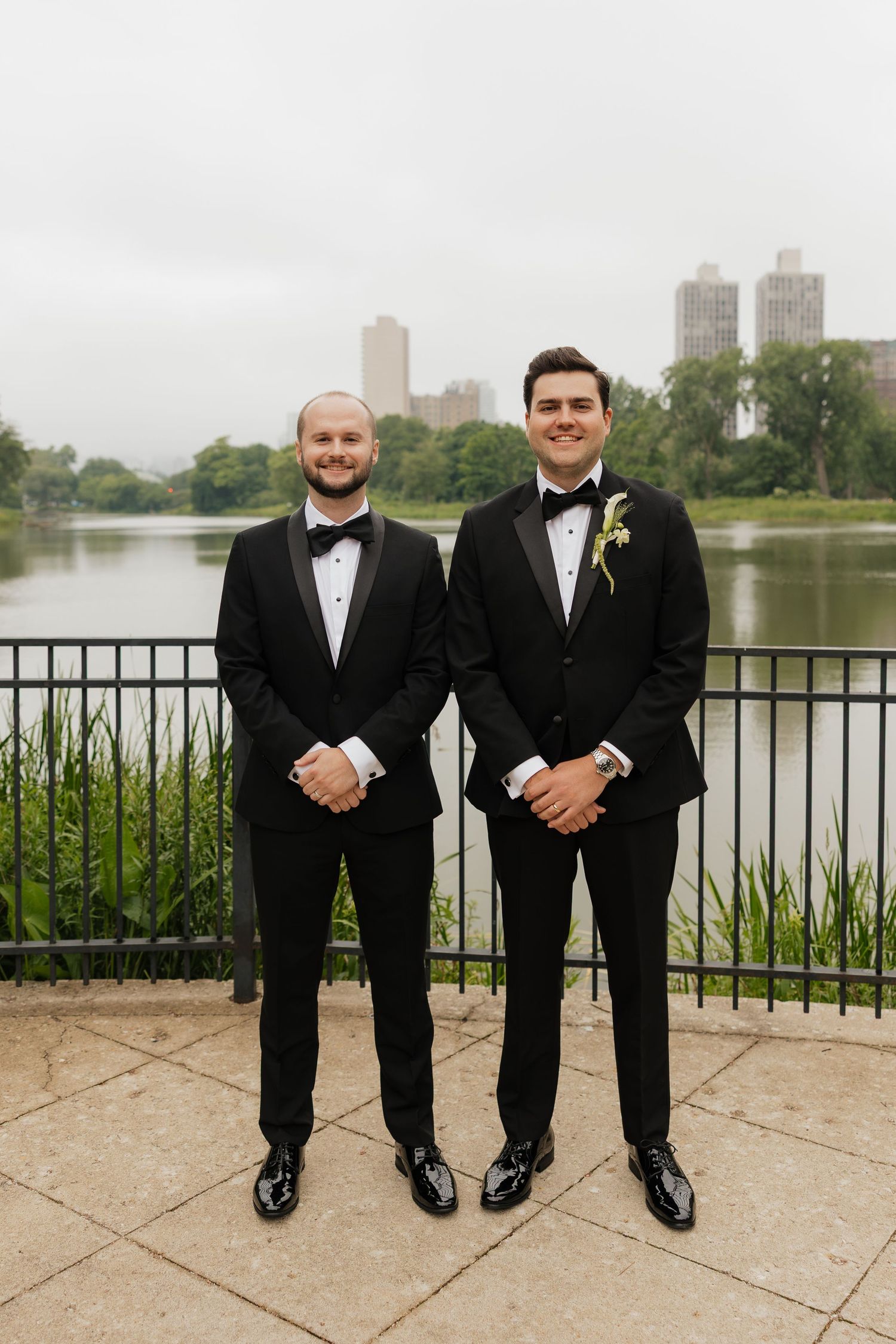 Two grooms in black tuxedos with boutonnieres pose by a lakeside railing with city skyline backdrop.