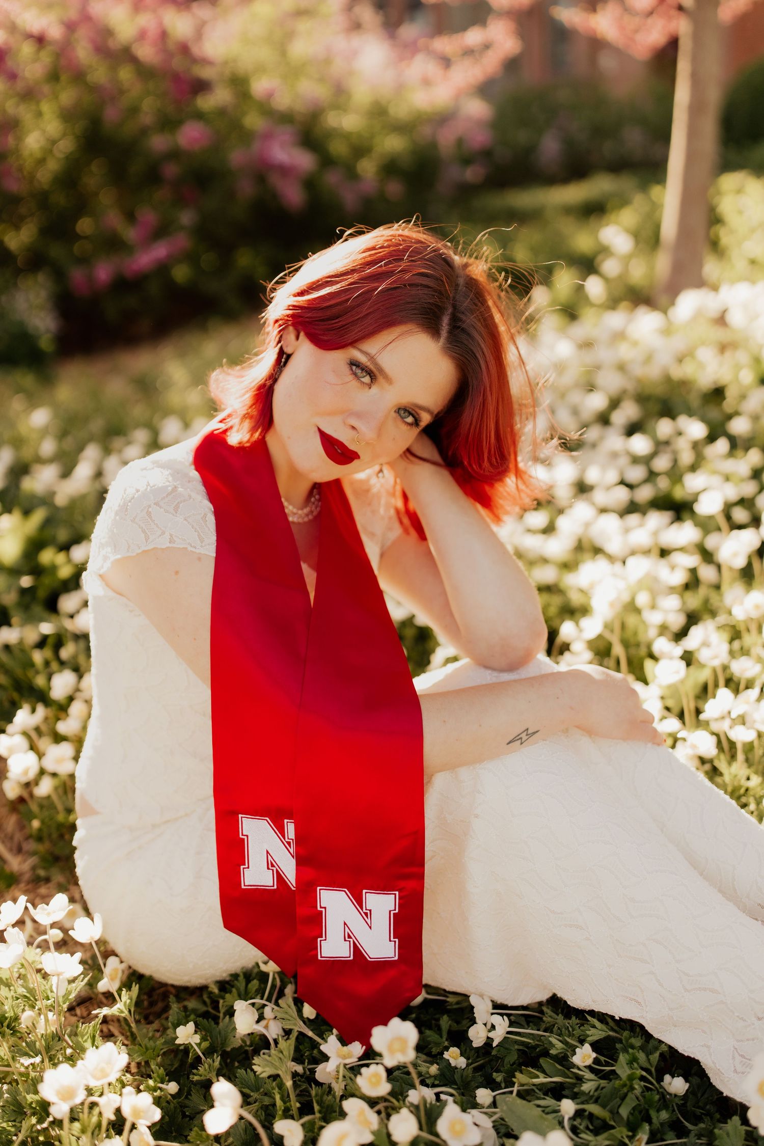 A person in a white dress and red Nebraska scarf sits among blooming flowers in a sunlit garden.