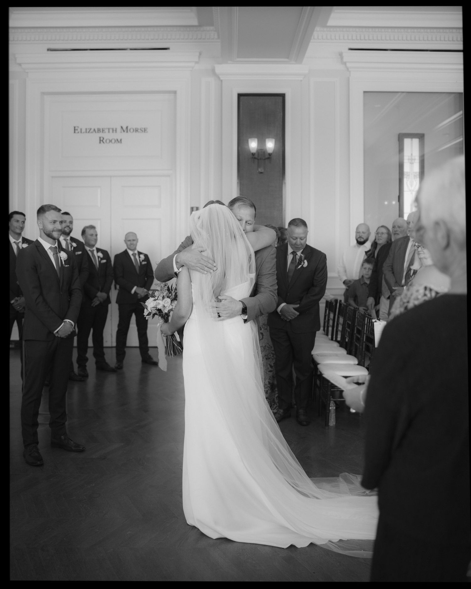 Black and white sequence of a wedding ceremony in a grand hall with guests looking on.
