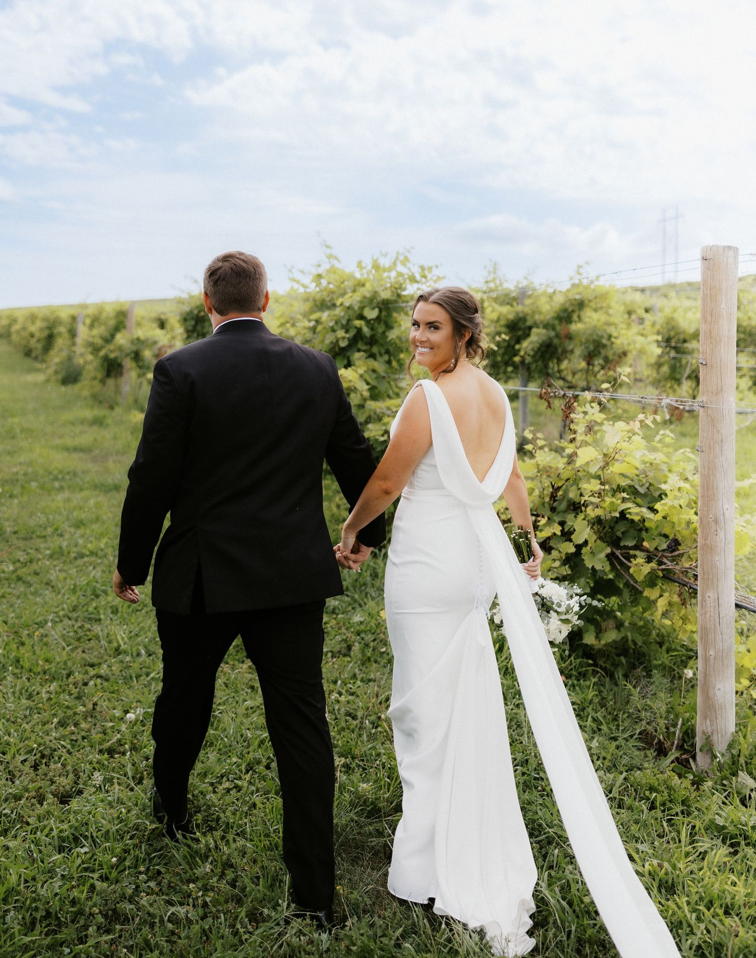 Bride and groom walk through lush green vineyard with flowing white dress and black suit on their wedding day.