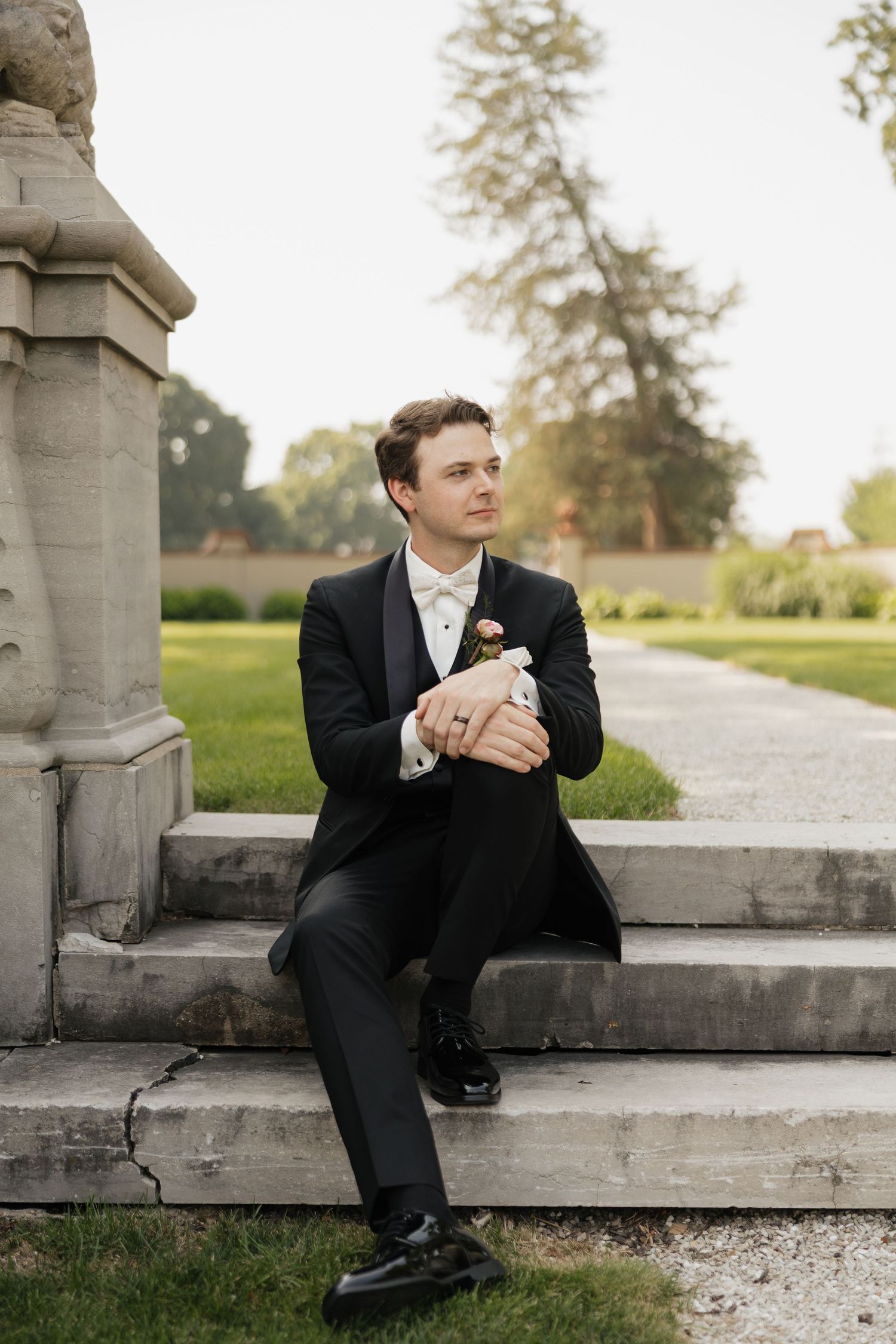 Groom in black tuxedo with boutonniere sits on stone steps in elegant garden setting.
