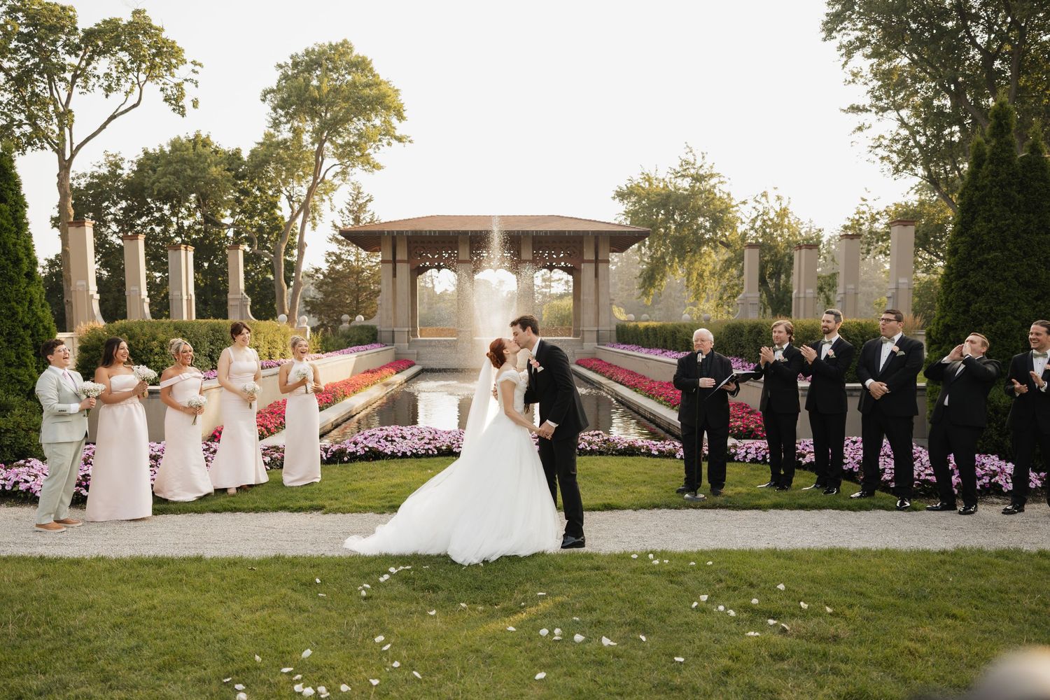 Bride and groom share a kiss surrounded by wedding party in a elegant garden venue with fountain backdrop.