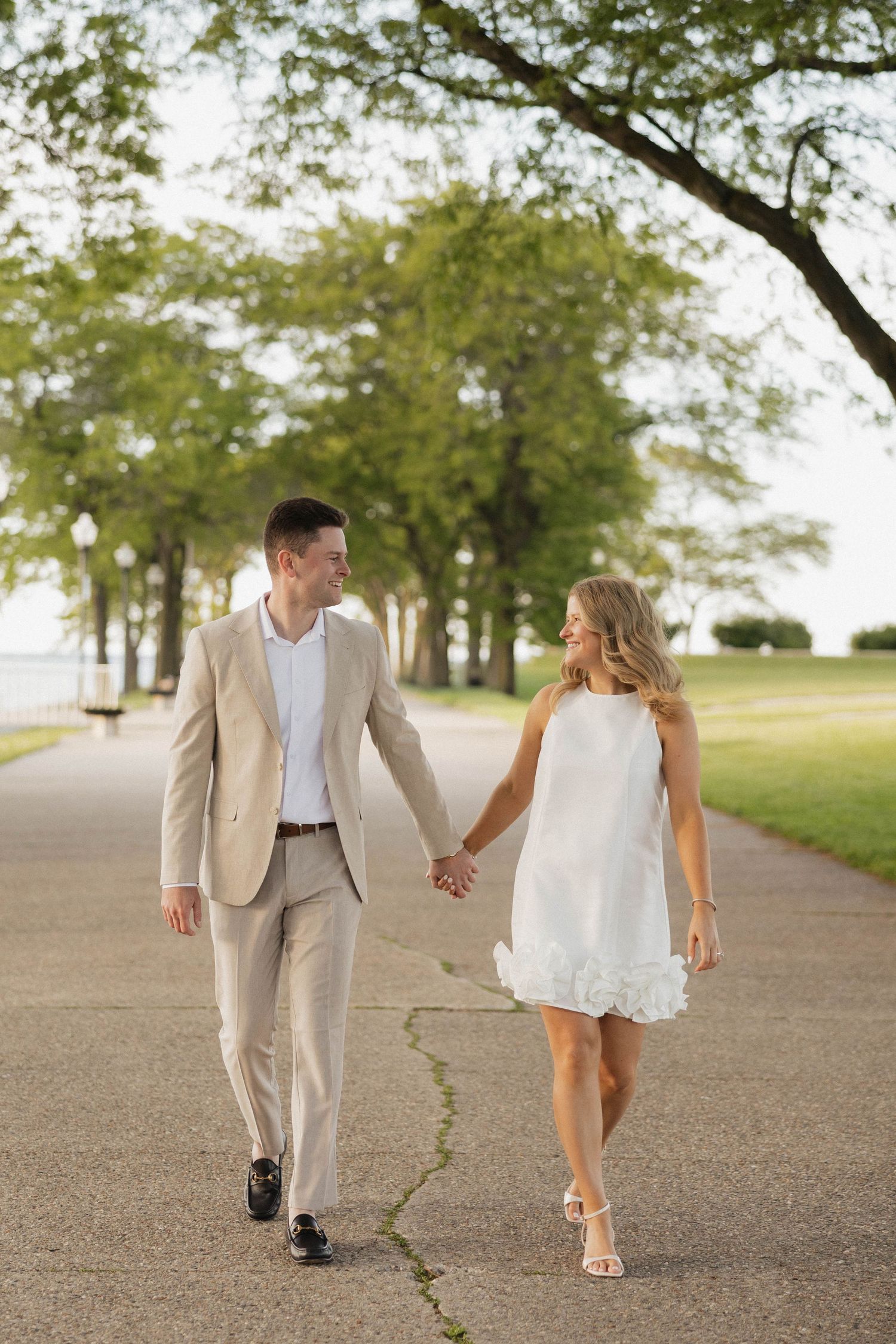 Couple holding hands walking on a tree-lined path, man in tan suit and woman in white mini dress.