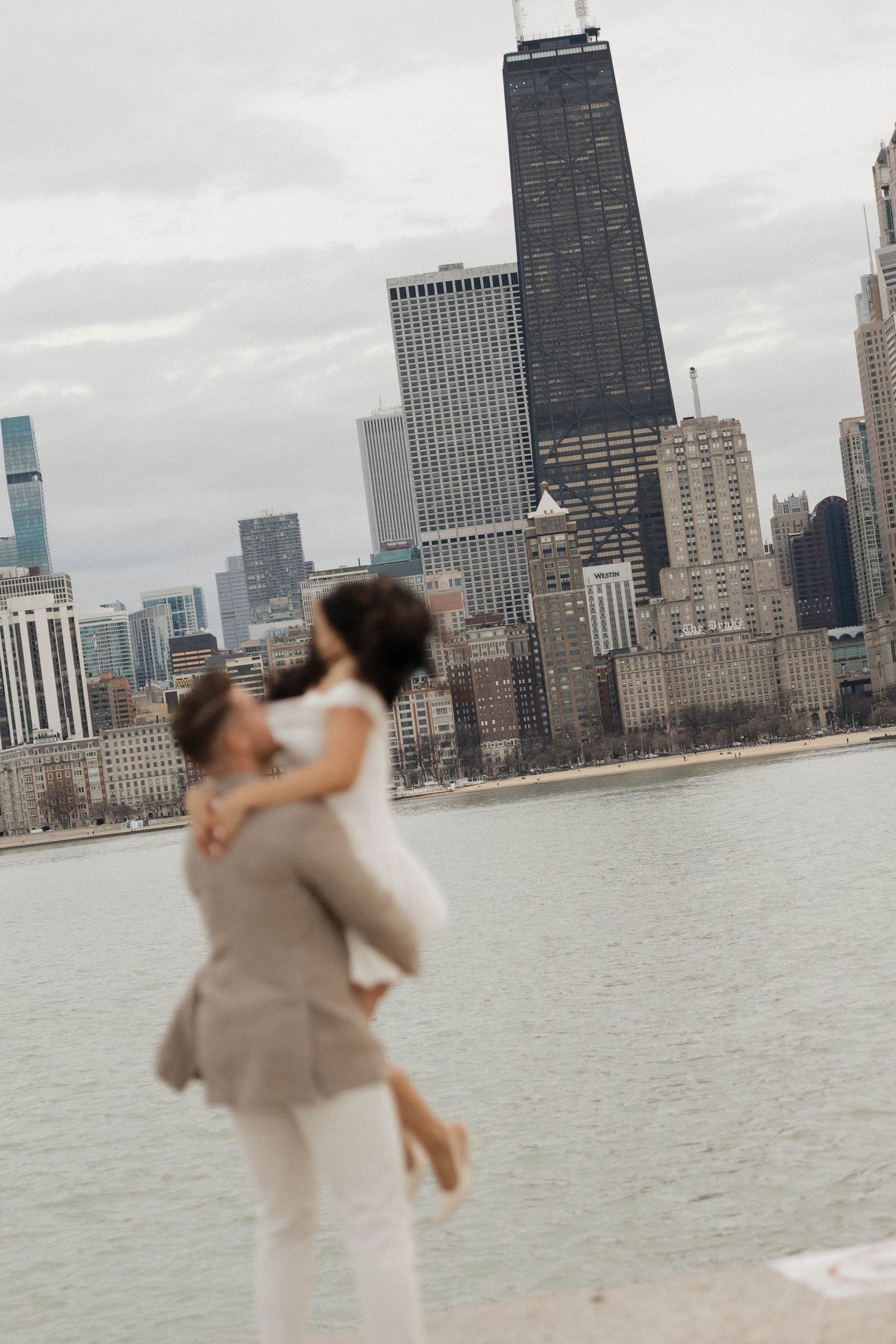 Couple embracing in front of Chicago skyline with John Hancock Center visible on a cloudy day.