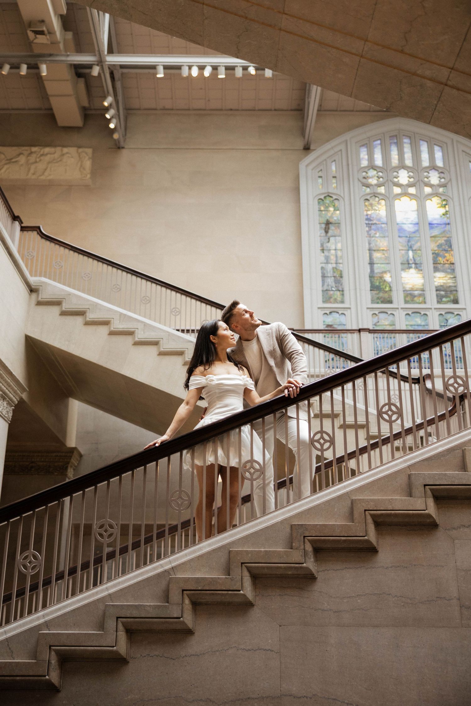 Romantic couple shares a tender moment on elegant marble staircase beneath a stunning stained glass window.