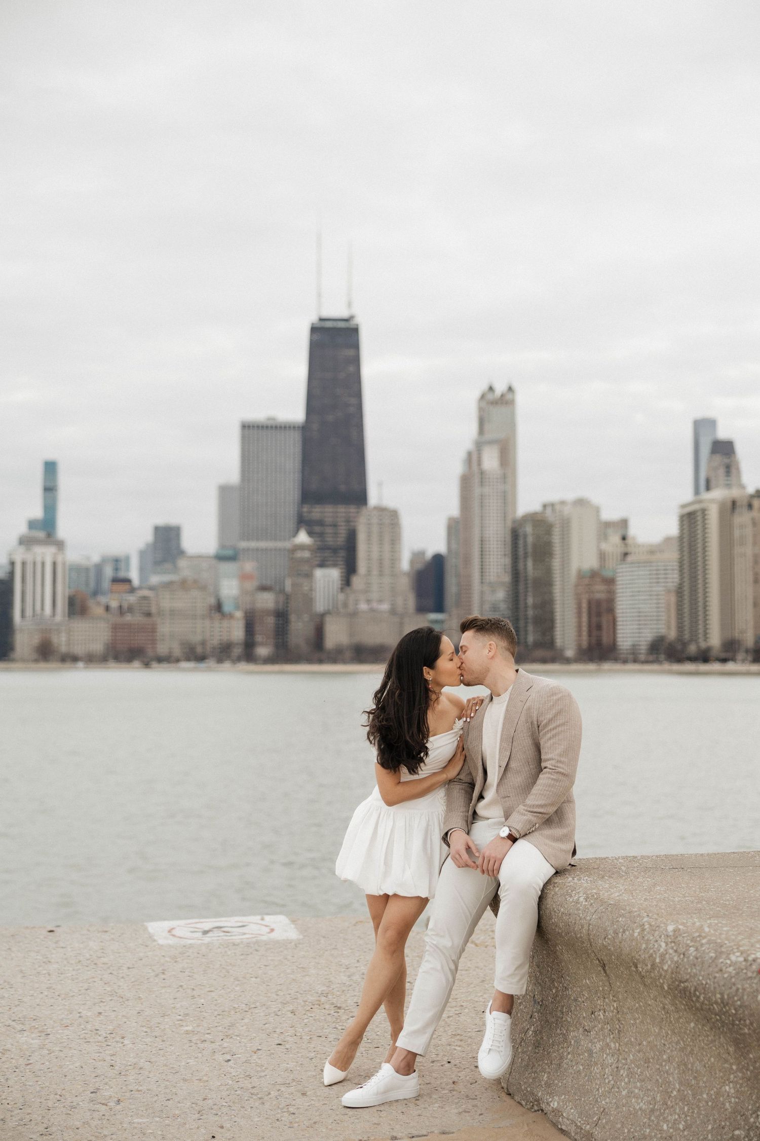 Couple sharing a romantic kiss on Chicago lakefront with iconic city skyline and John Hancock Center in background.