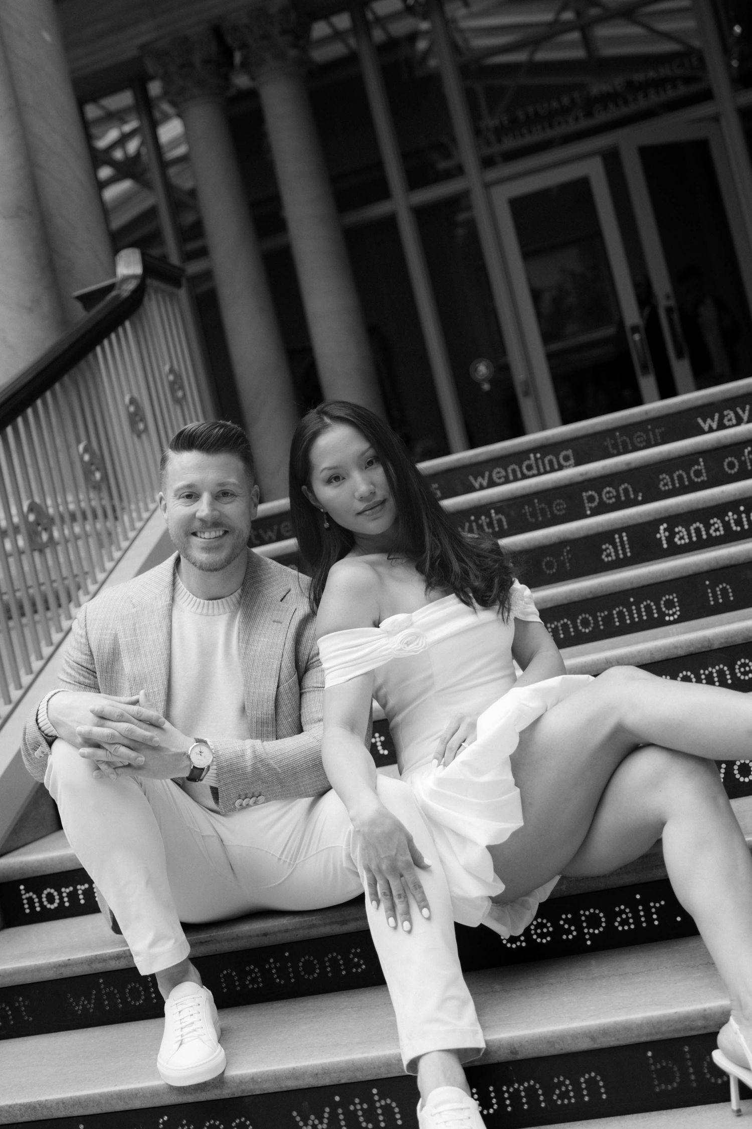 Black and white photo of a stylish couple sitting on steps with text inscriptions, woman in white dress, man in light suit.
