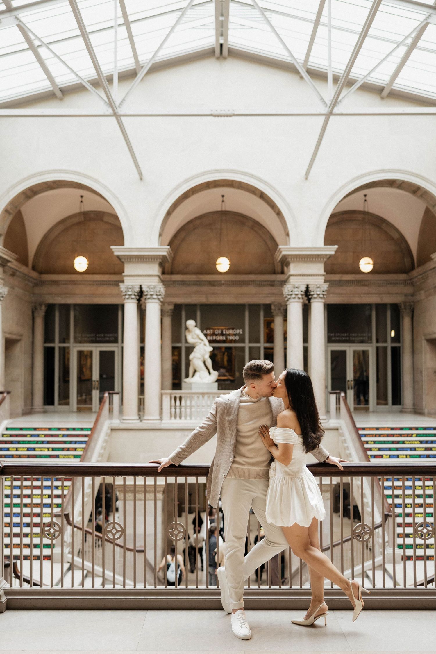 Couple kissing on a balcony inside an elegant museum atrium with arches and classical sculpture.