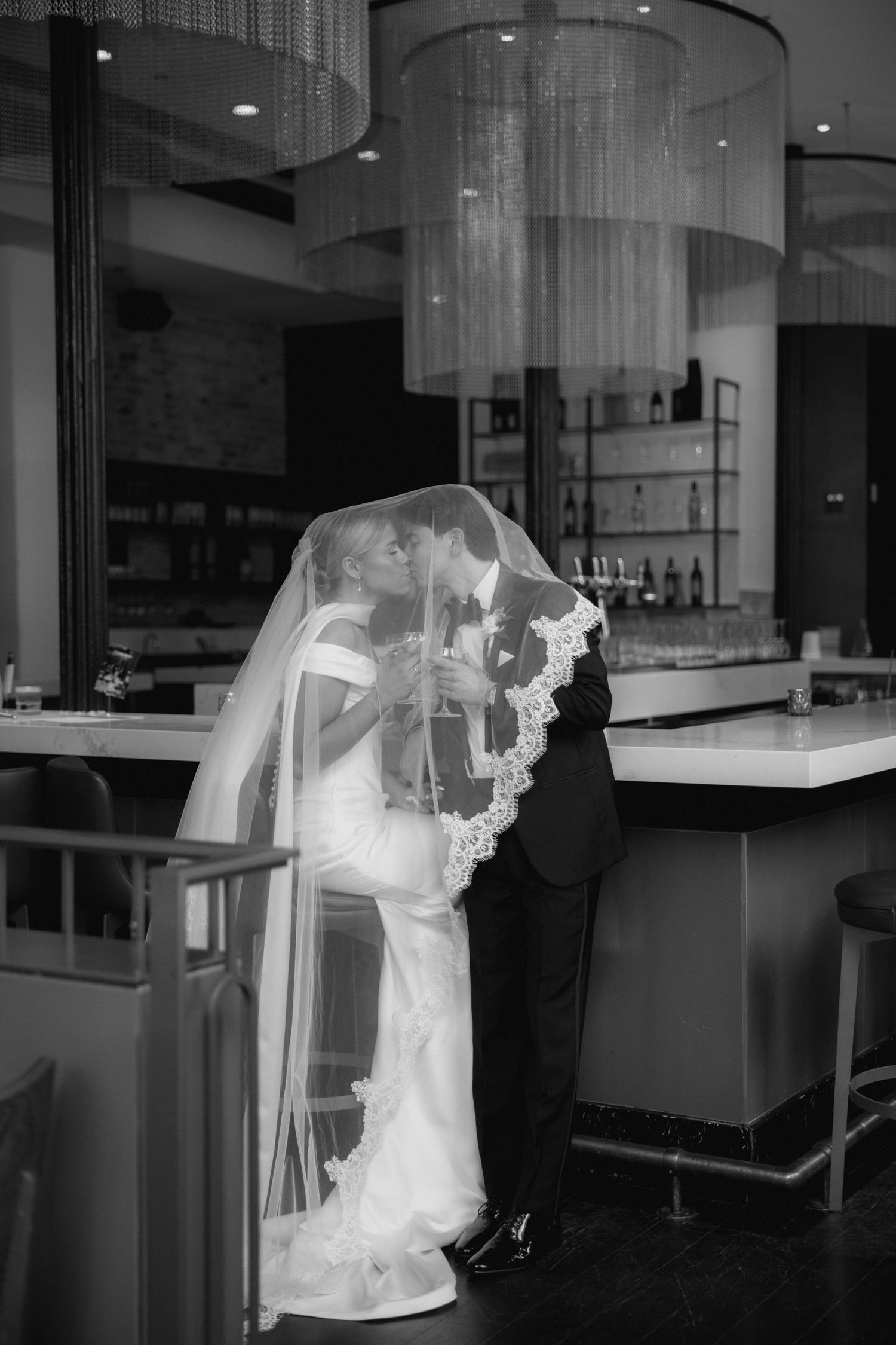 Black and white photo of bride and groom sharing a kiss under veil at elegant bar venue.