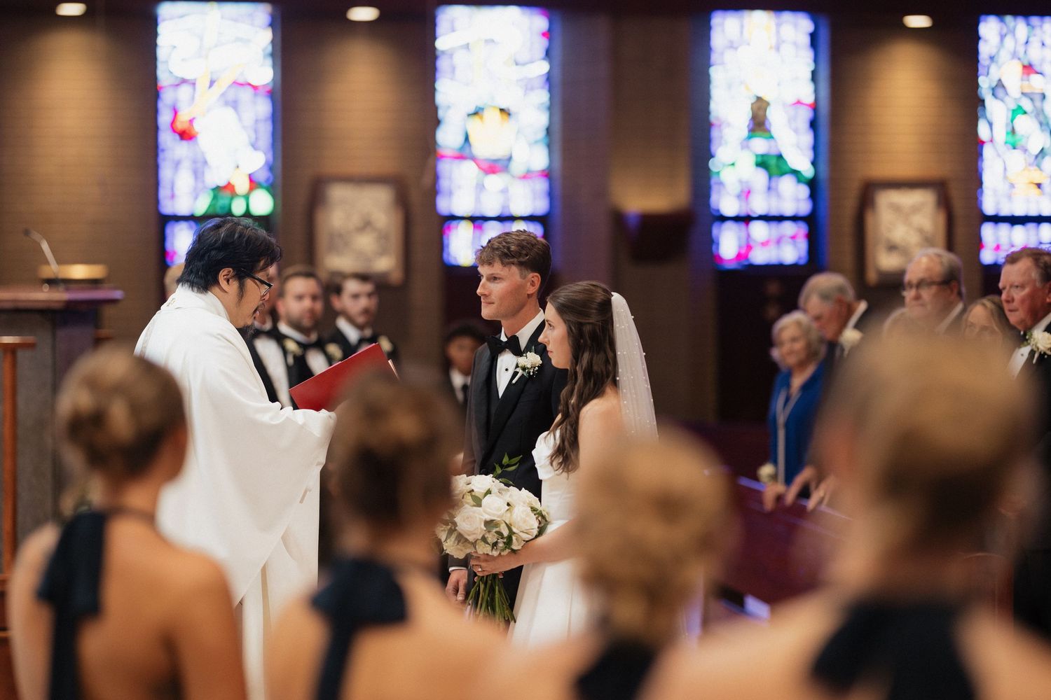 Bride and groom exchange vows at church altar with stained glass windows and wedding guests seated in pews.