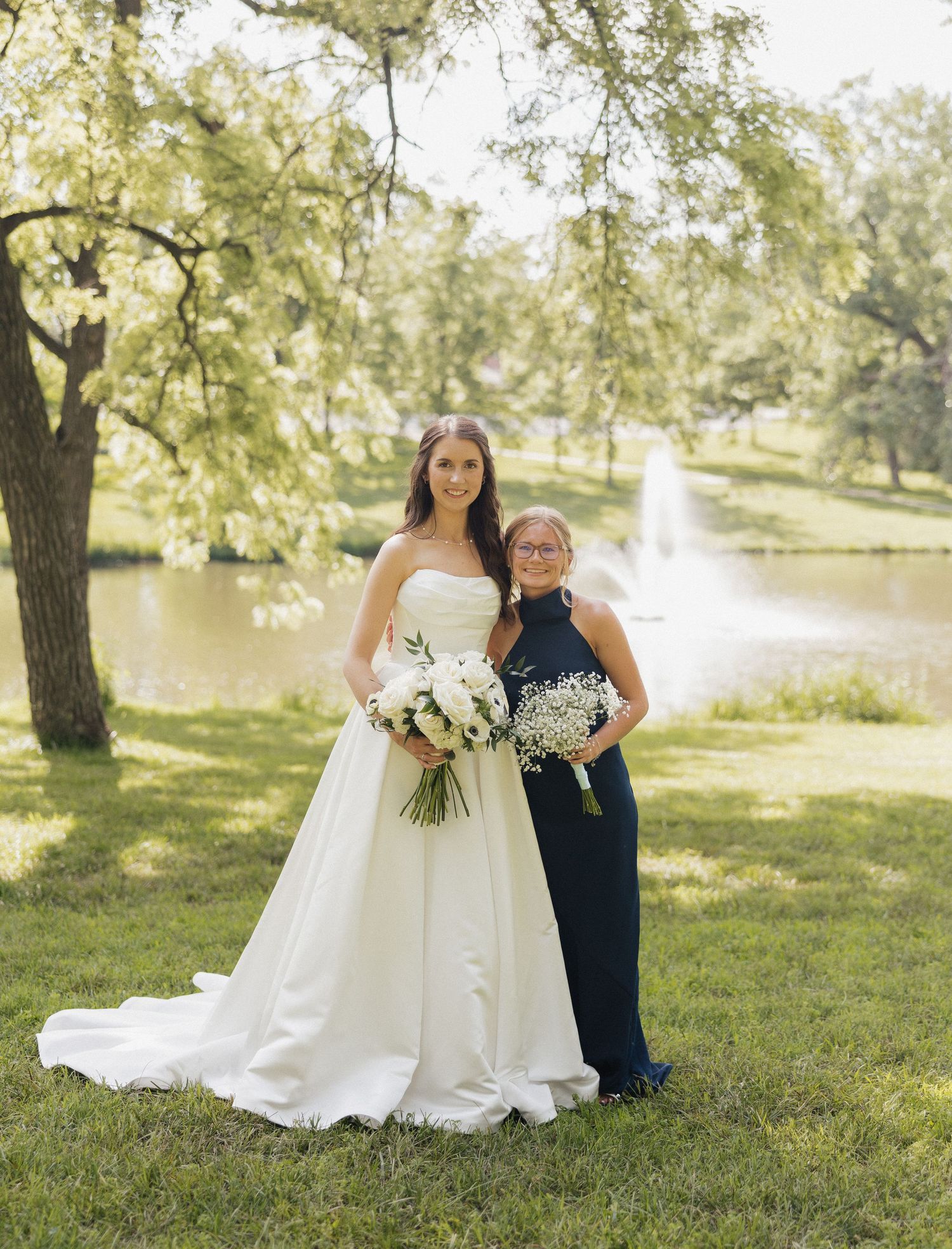 Bride in white strapless gown holds bouquet beside bridesmaid in navy dress near pond with fountain.