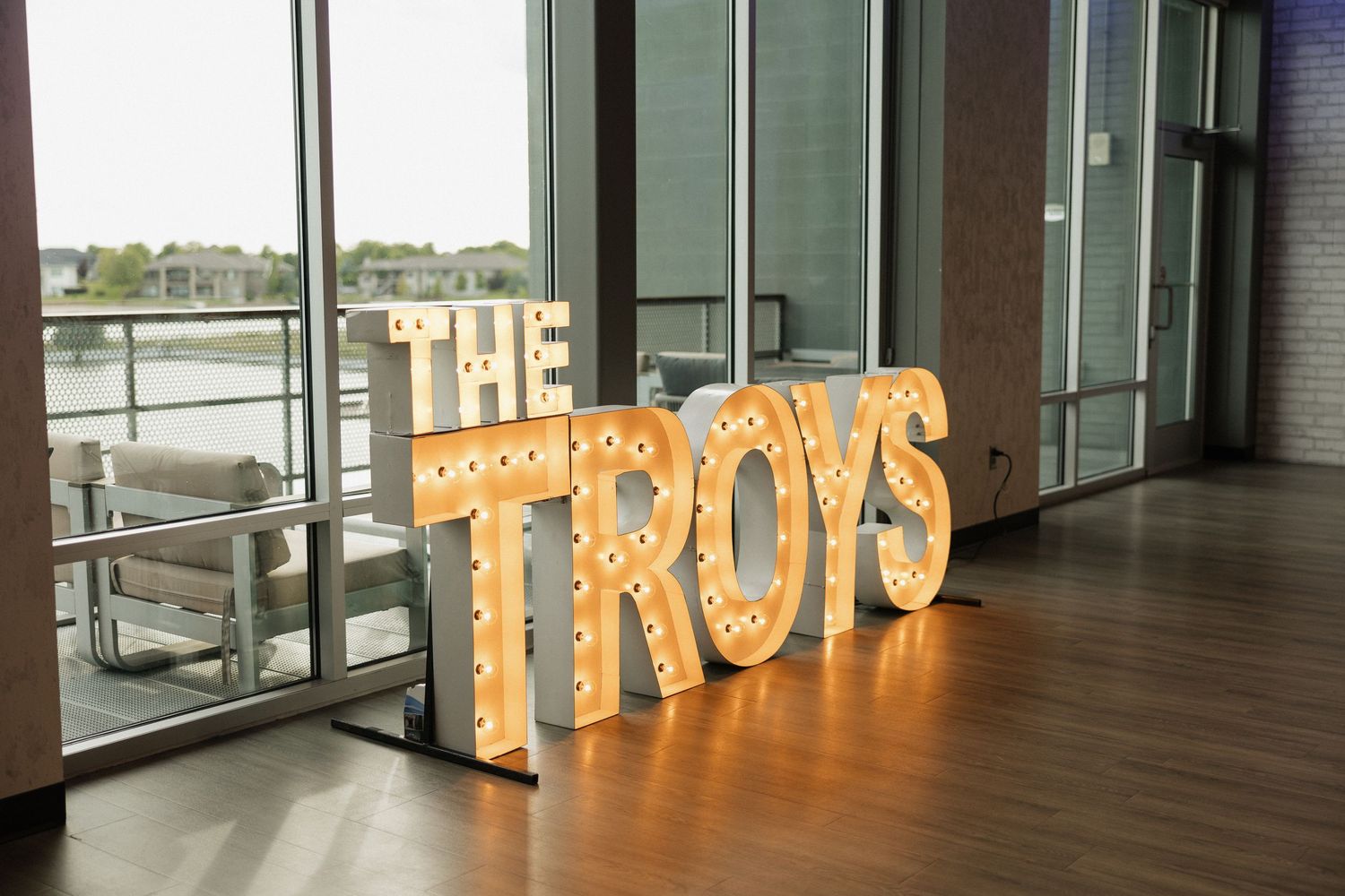 Illuminated marquee letter sign spelling THE TROYS with light bulbs, displayed indoors near large windows.