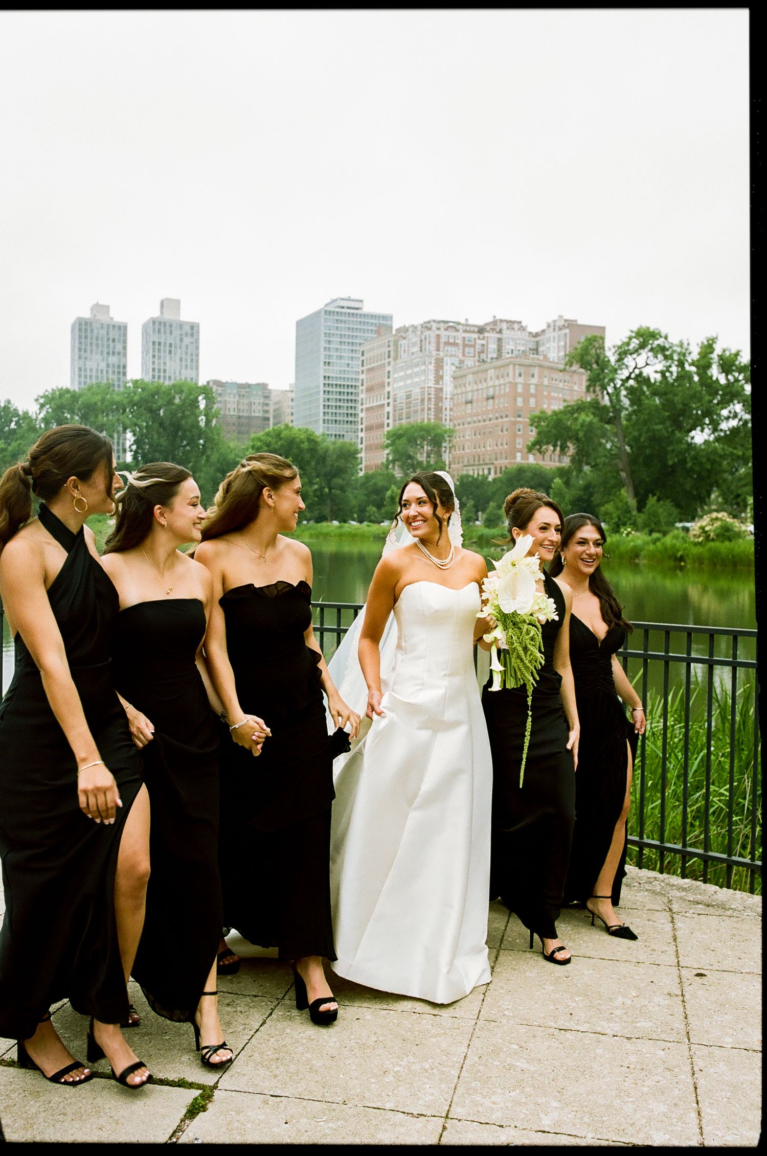 Bride in white gown walks with bridesmaids in black dresses near Central Park lake with NYC skyline backdrop.