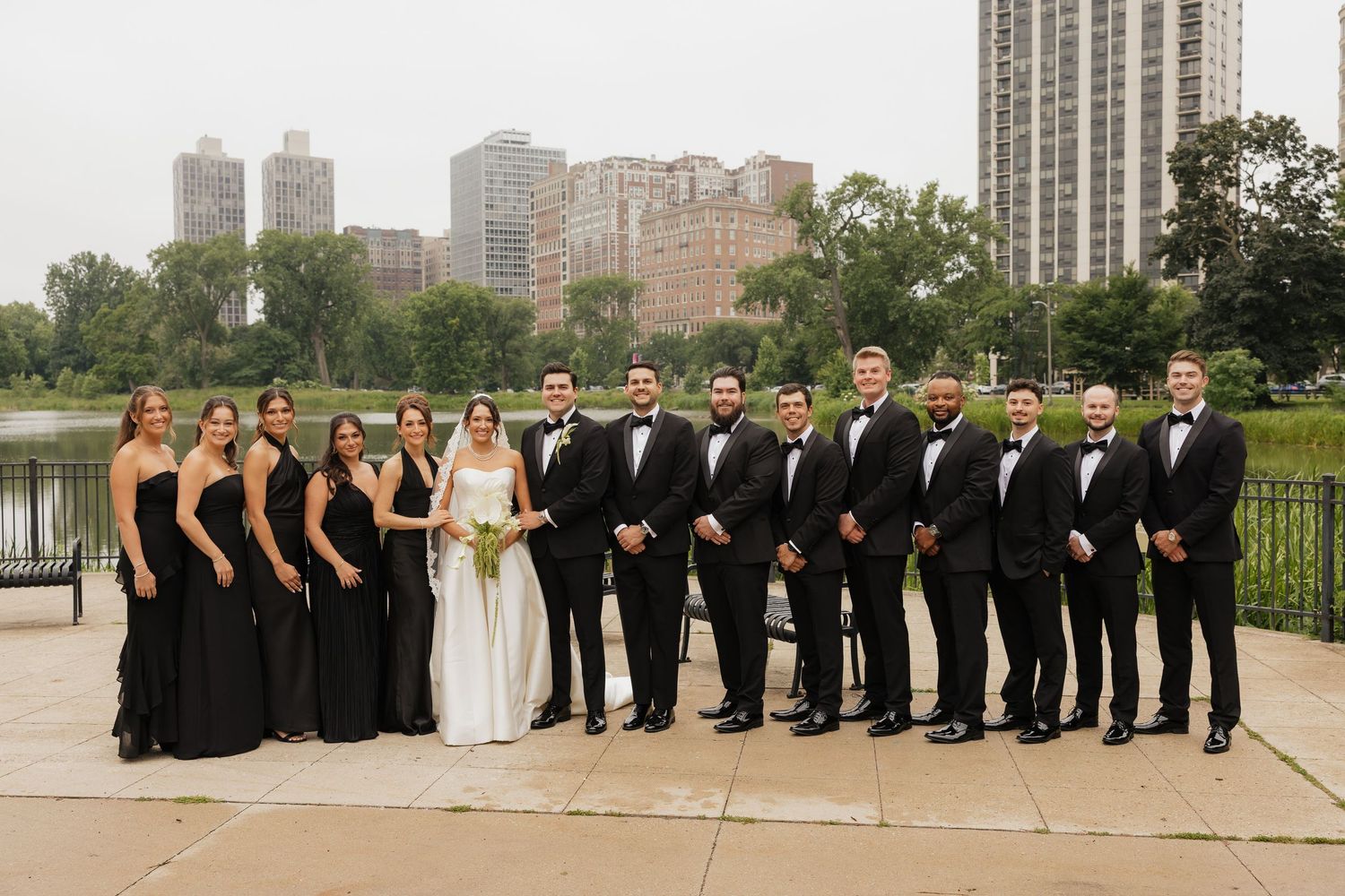 Wedding party in black attire poses outdoors near a lake with city skyline backdrop on a cloudy day.