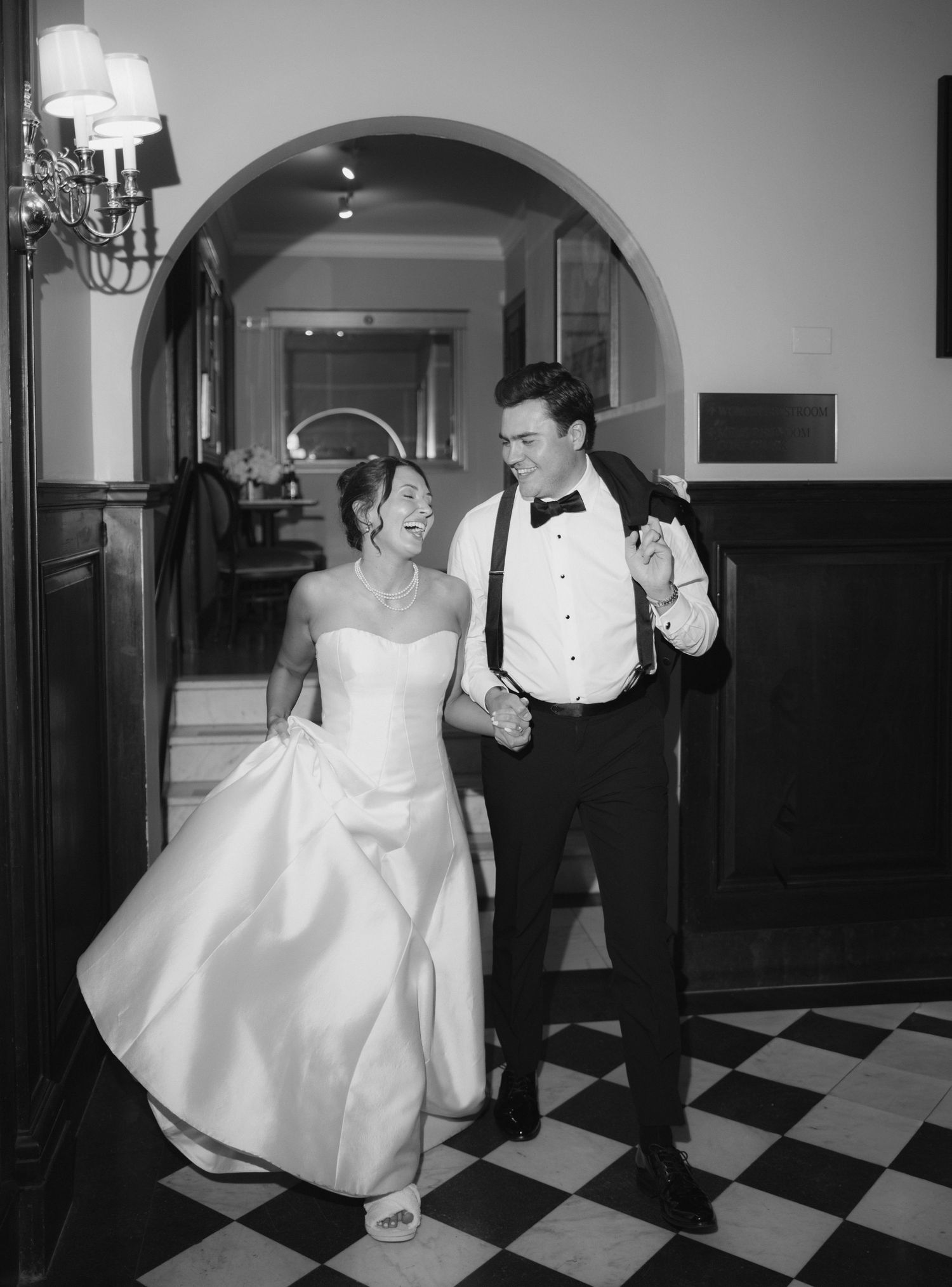 Black and white photo of a smiling bride in strapless gown and groom in tuxedo walking through arched doorway.