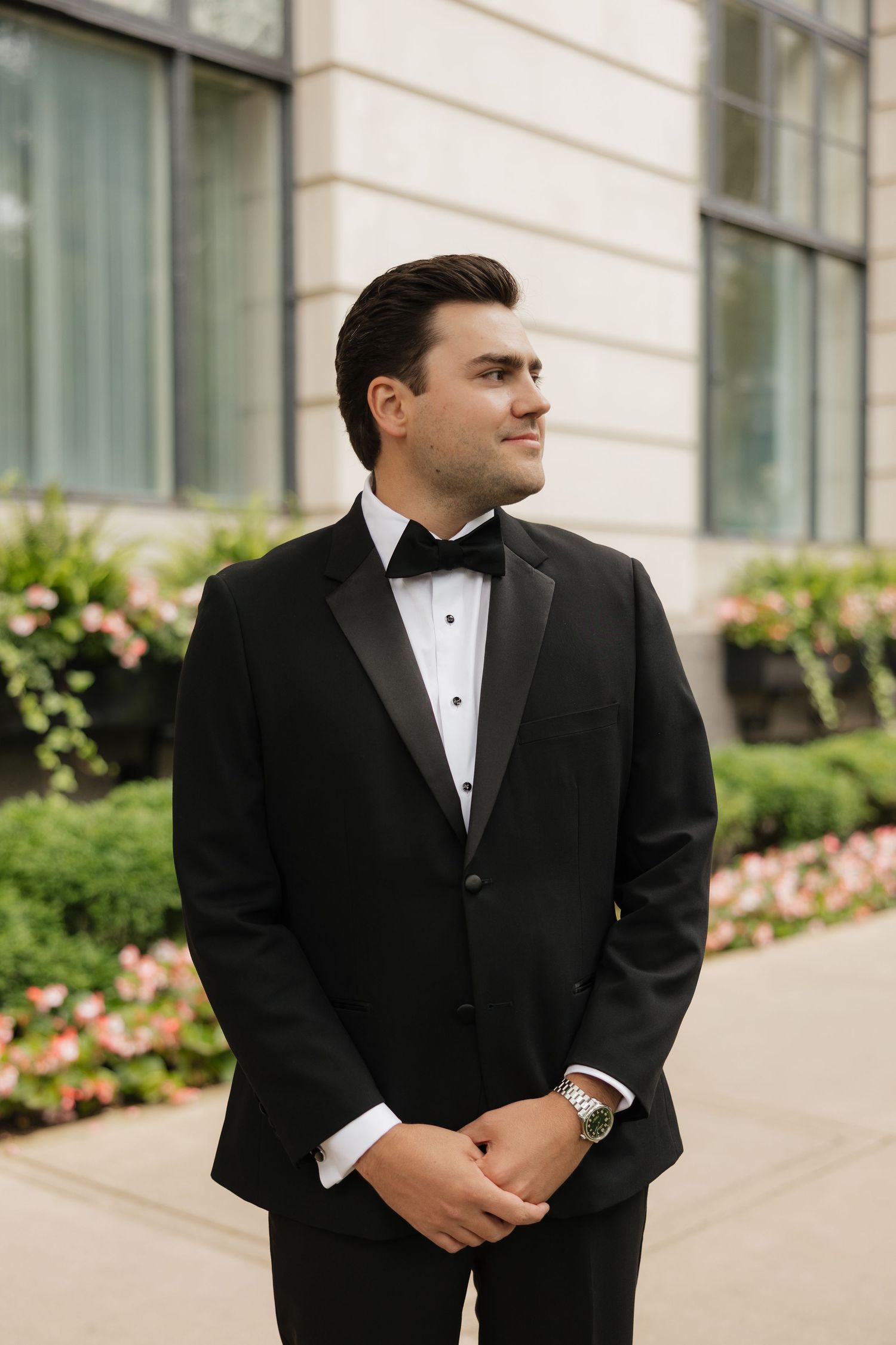 A dapper groom in a black tuxedo with bow tie and watch stands outdoors near flowering bushes.