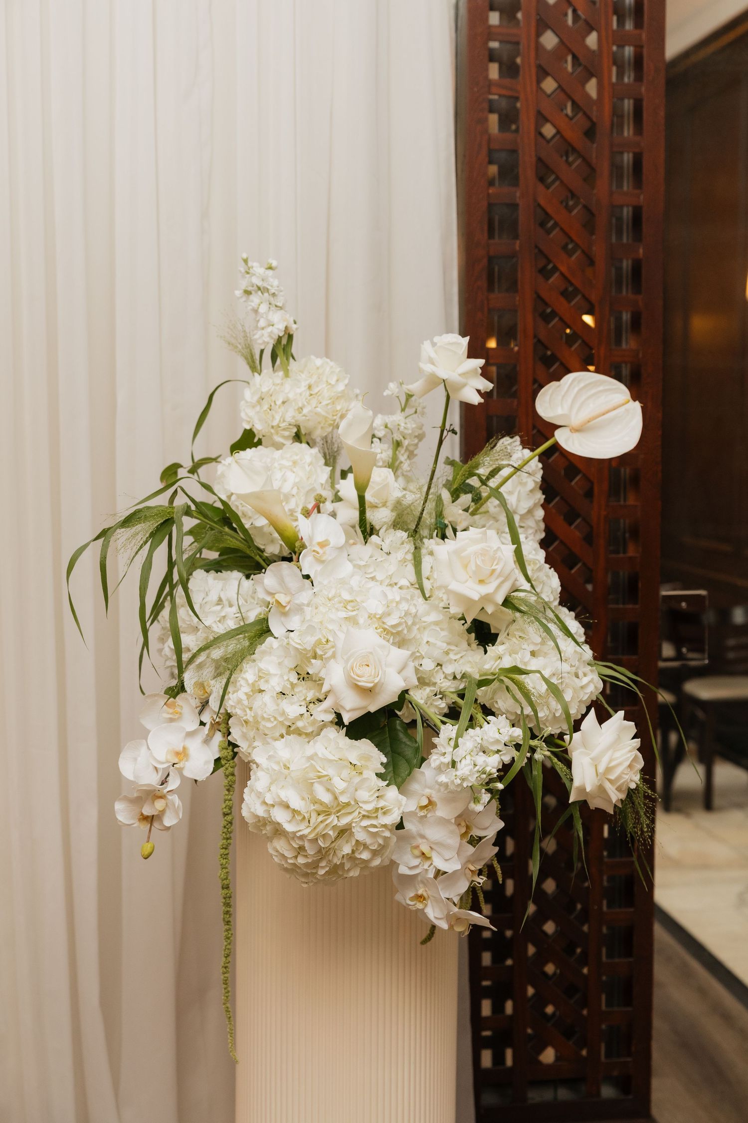 Elegant white floral arrangement with hydrangeas, roses, orchids, and anthurium in a white vase against draped curtains.