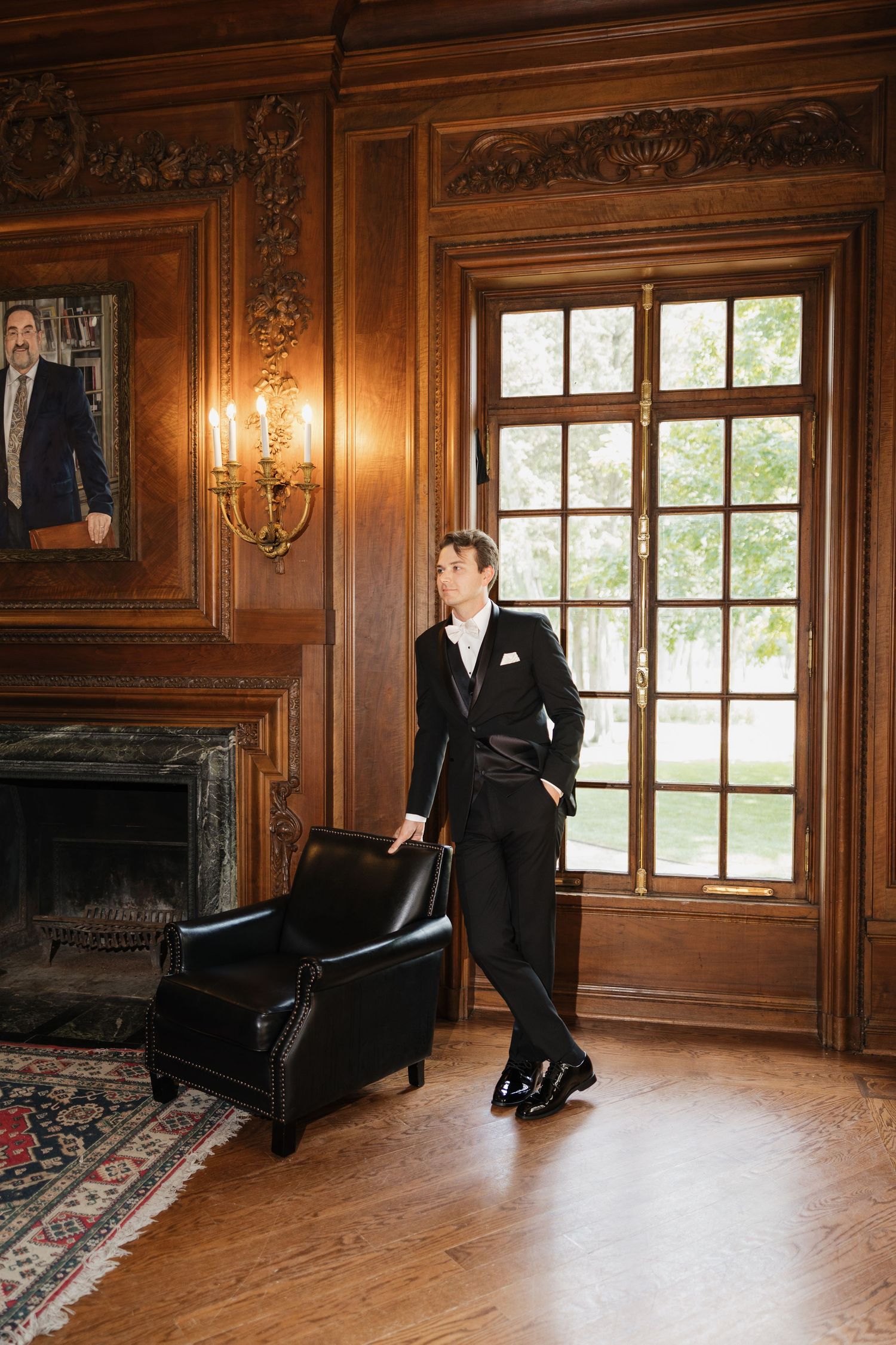 A groom in a black tuxedo stands by a leather chair near a large window in an elegant wood-paneled room with a fireplace.