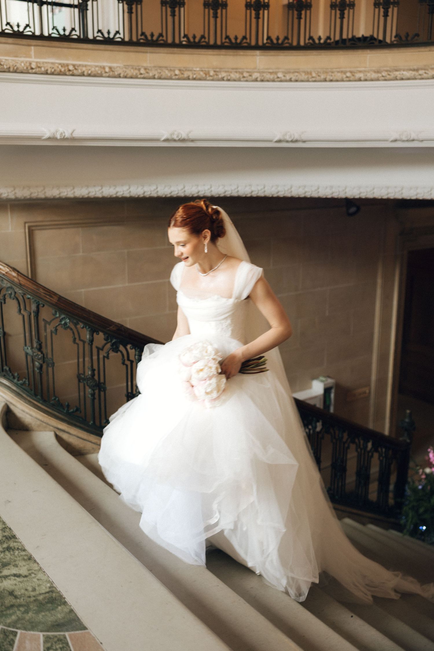 Radiant redhead bride in layered white ball gown descends grand staircase holding white peony bouquet.