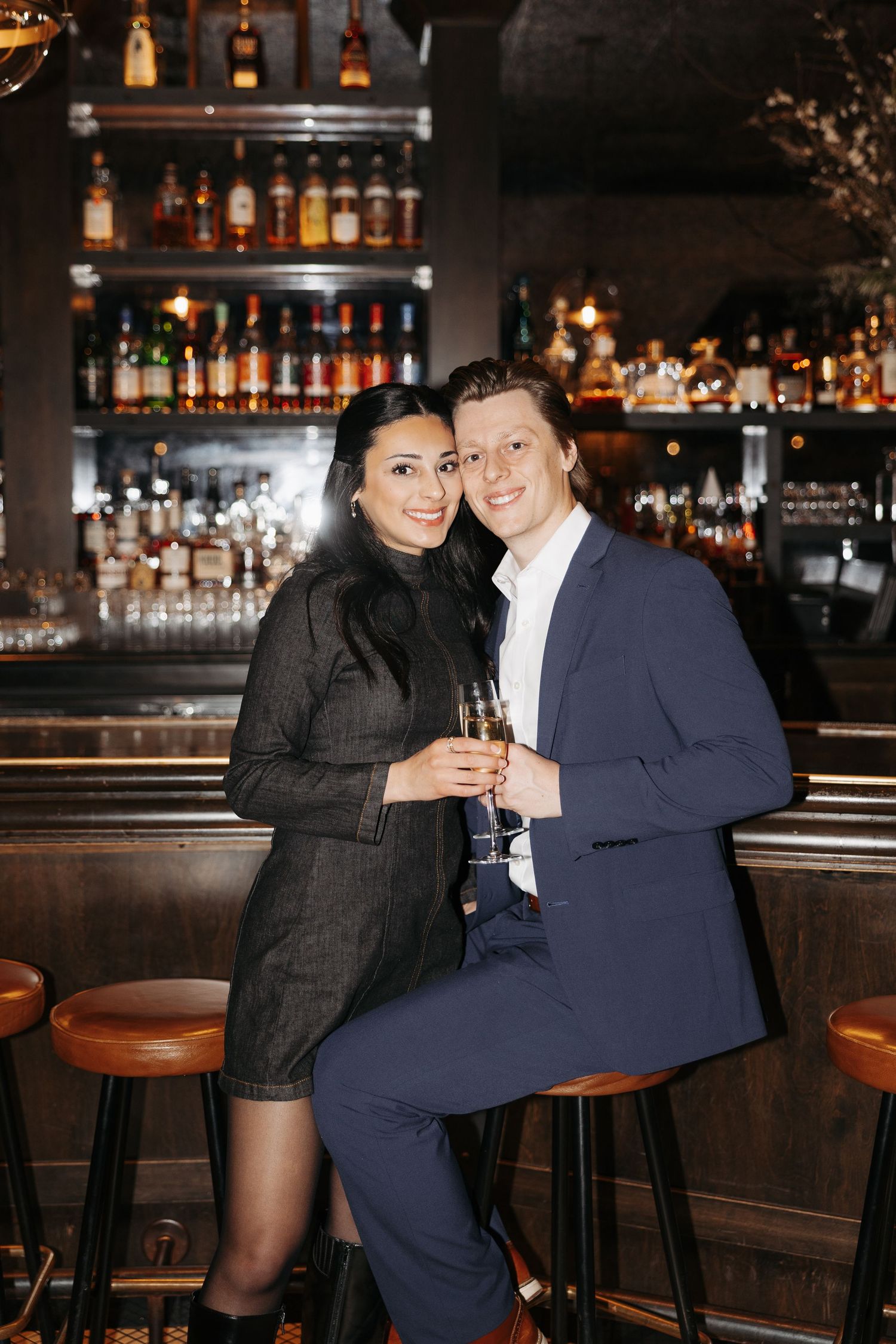 Stylish couple sharing drinks at a dimly lit upscale bar with bottles displayed in background.