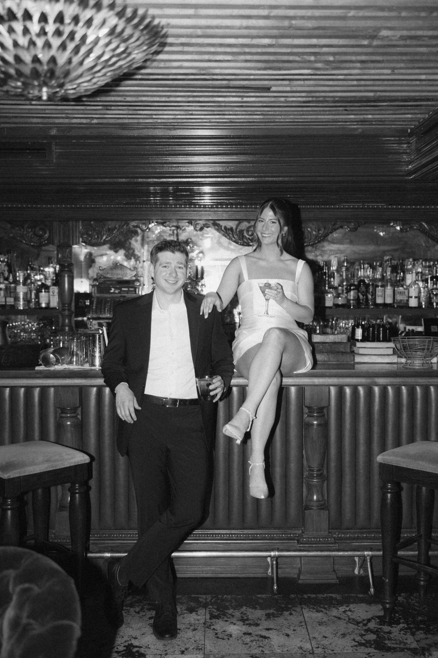 Black and white photo of two people posing at a vintage bar counter with stools and bottles in the background.