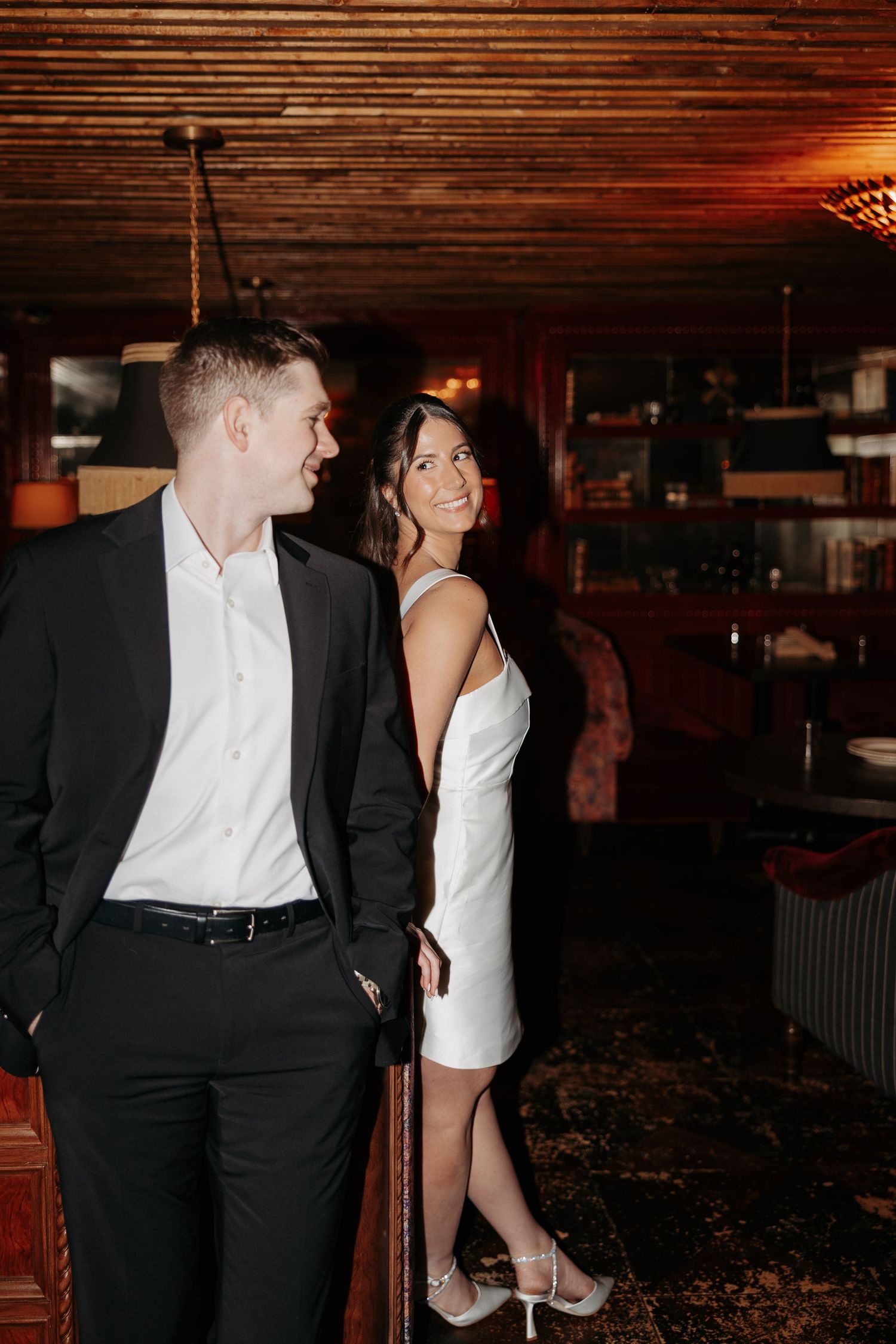 A couple in formal black and white attire pose together in a dimly lit indoor setting.