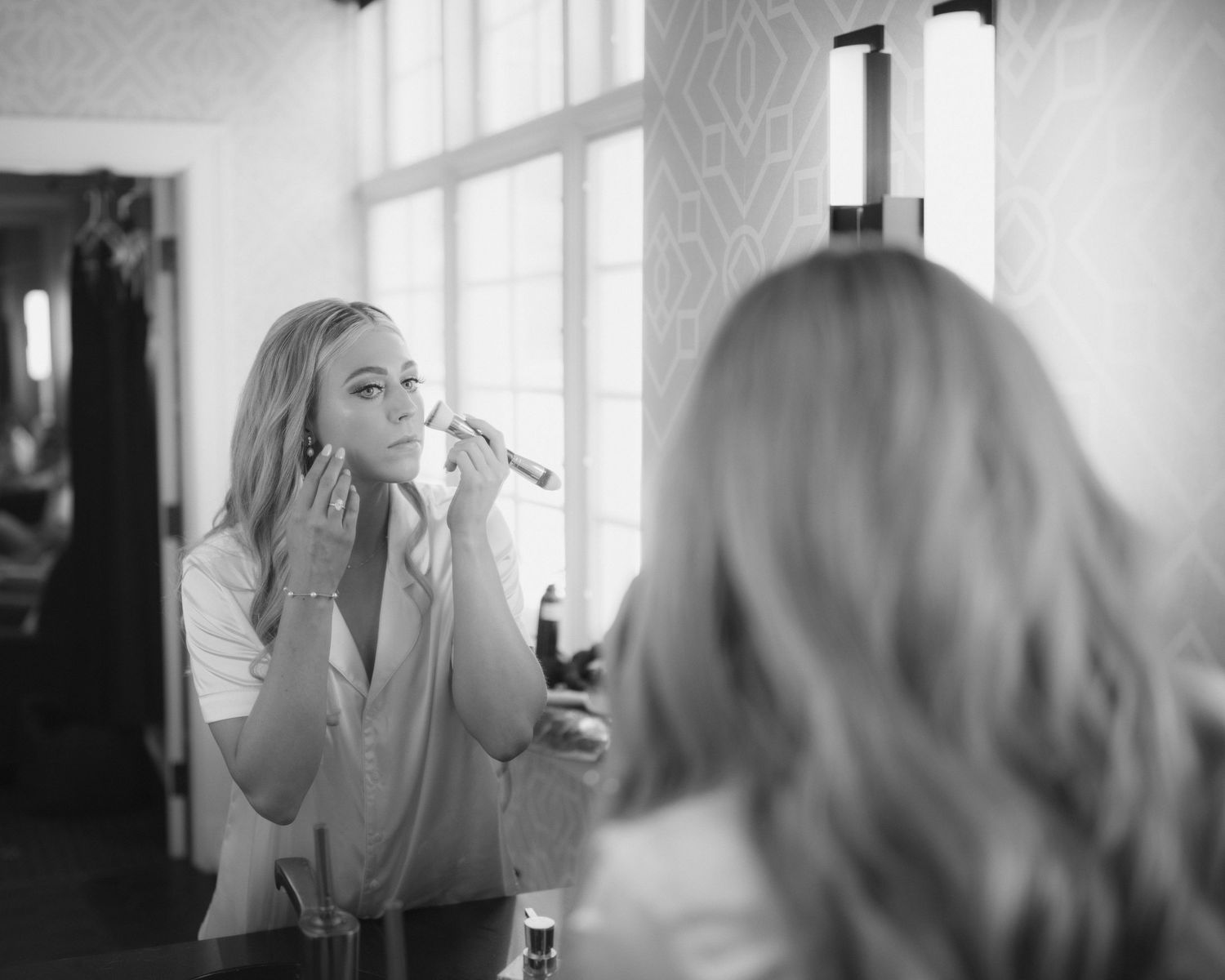 A series of black and white photos showing someone applying makeup in a mirror with soft natural lighting.