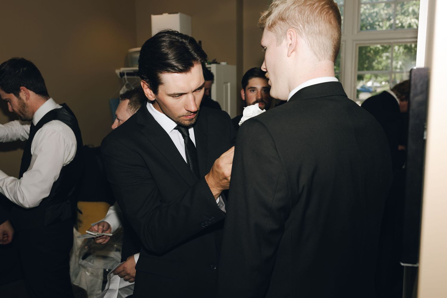 Two people in black suits adjust their formal attire while getting ready at an indoor event.