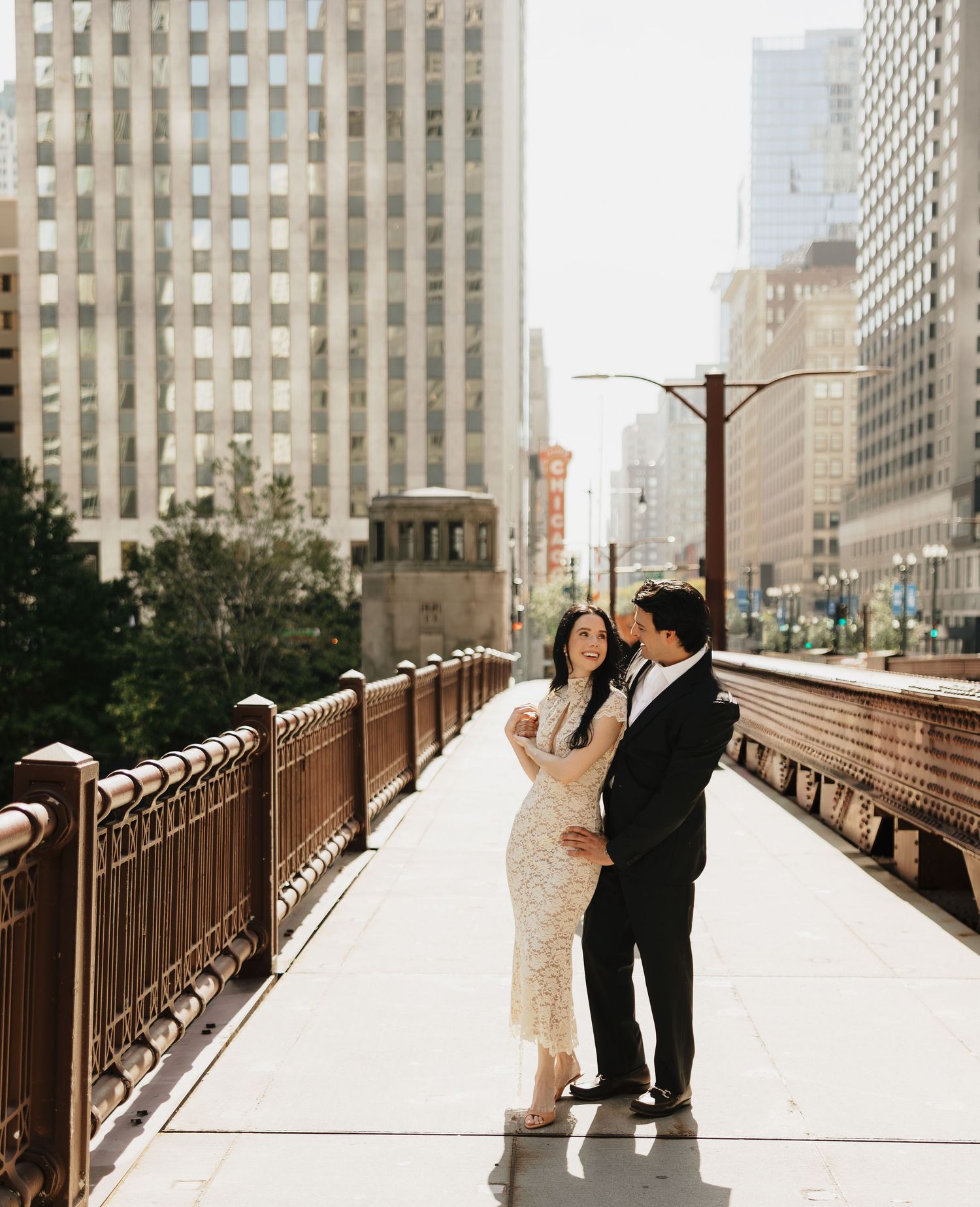 Couple in formal wedding attire embracing on a sunlit city bridge with ornate railings and downtown skyscrapers behind them.