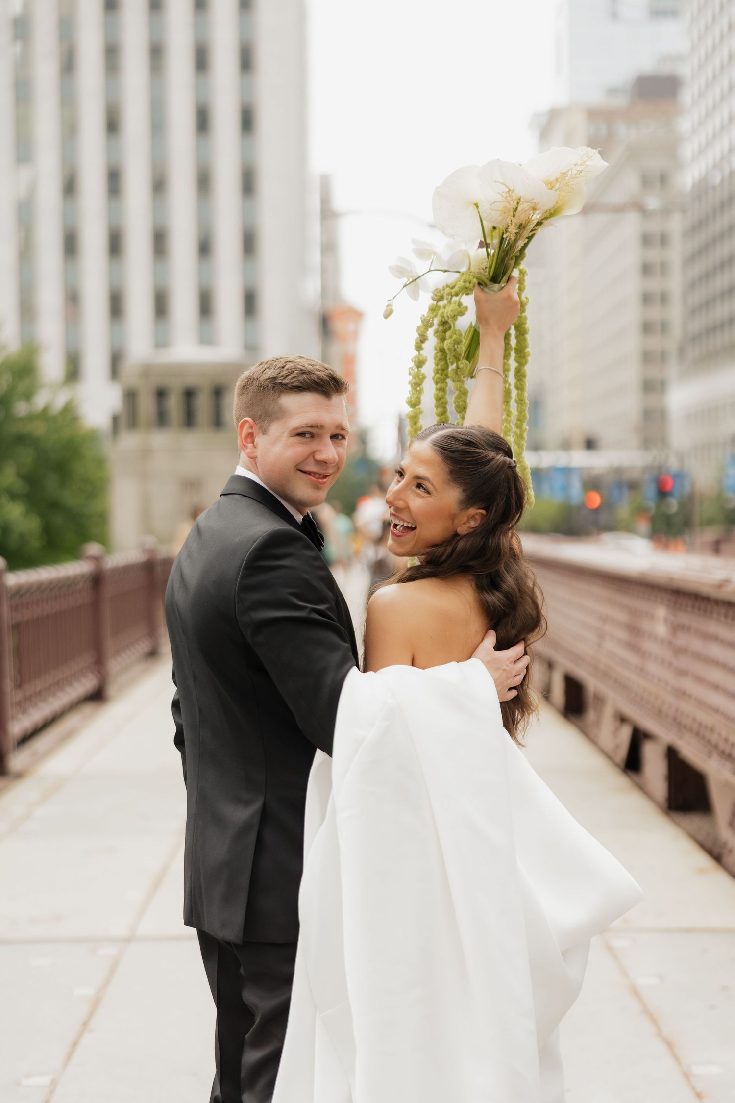 Smiling bride in a white gown raises her bouquet of white flowers and greenery as she walks with her groom in a suit on a city bridge.