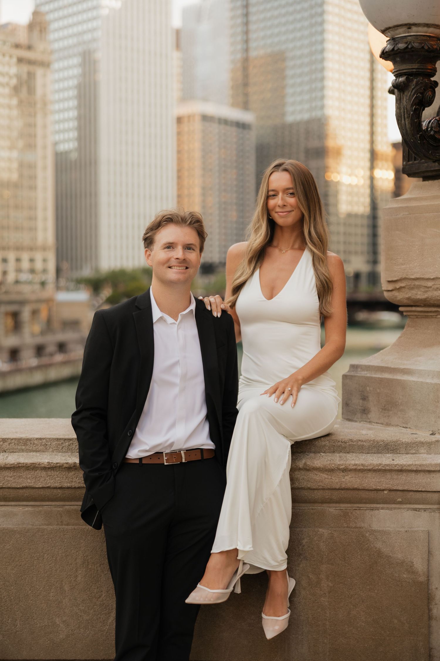 Smiling couple in elegant attire pose on a stone ledge with a river and downtown city skyscrapers behind them.